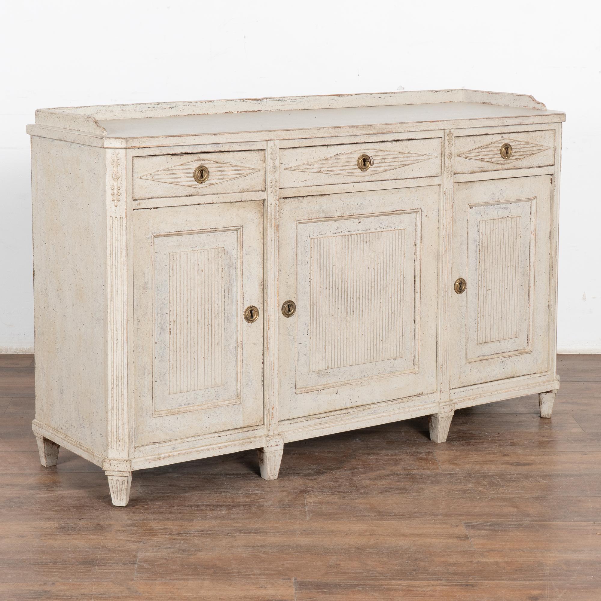 Gustavian white painted pine sideboard or console with one interior shelf.
Traditional fluted panel doors, fluted canted sides and fluted diamond accent along 3 upper drawers, raised on tapered fluted feet.
The newer, professionally applied antique