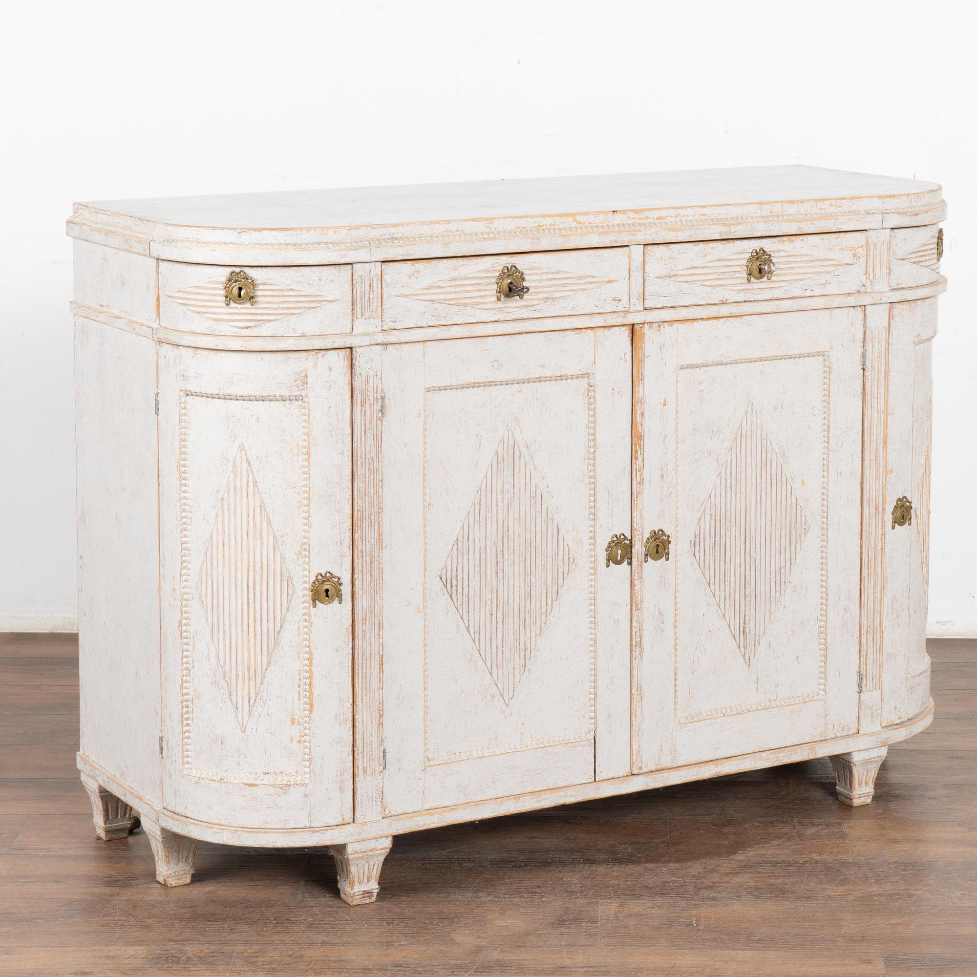 Swedish Gustavian white pine sideboard with attractive curved sides and traditional carved fluted diamond details.
The curved end cabinet doors and drawers make this cabinet a special find.
The newer professionally applied soft antique white painted