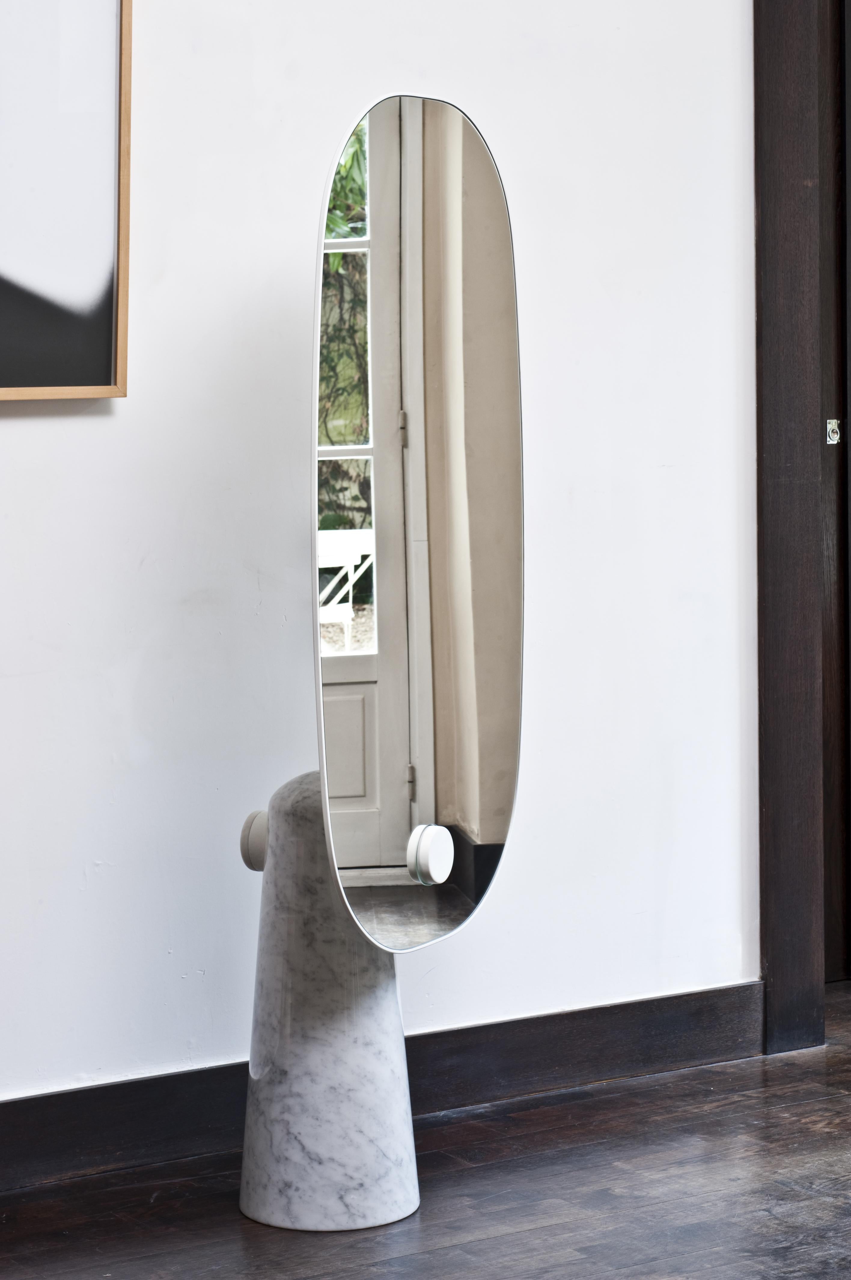 Iconic is a mirror that proudly stands as a statue. The white or black marble base pedestal supports a thin
metal structure.
The mirror seems to float above the base while an horizontal metal cylinder intersects both parts.
This oversized, free