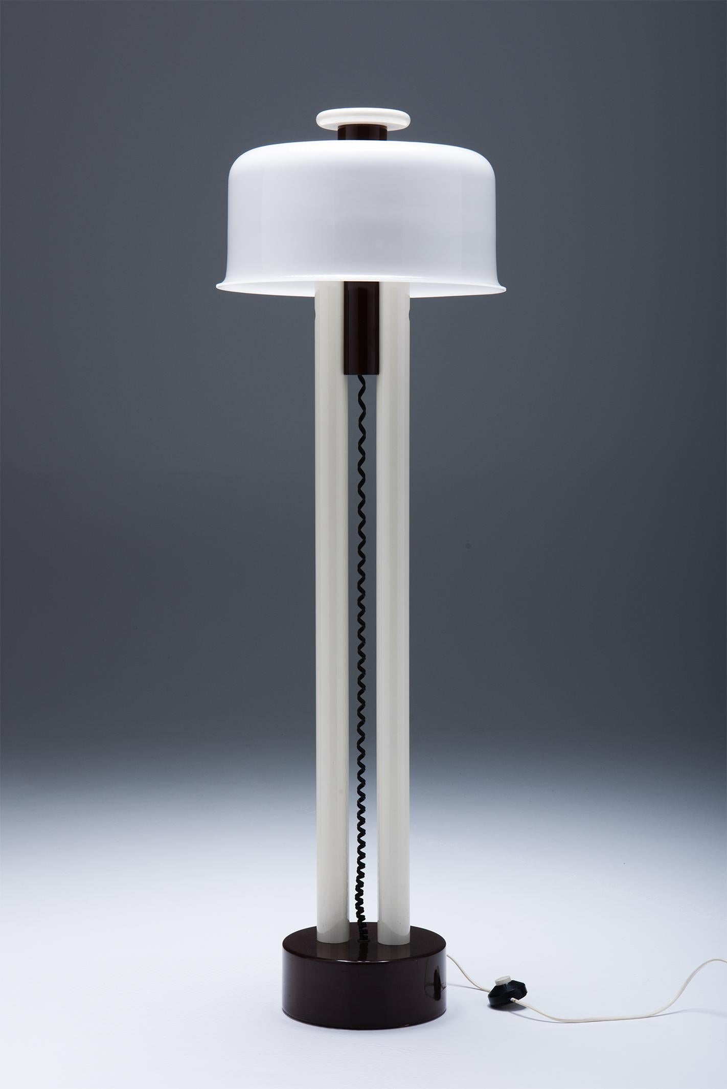 Aquí tenemos una lámpara de pie postmoderna italiana.
La estructura consta de dos pilares, fabricados en metal pintado de blanco. Entre estos dos pilares podemos ver un alambre. La lámpara descansa sobre una base de madera lacada en marrón y tiene