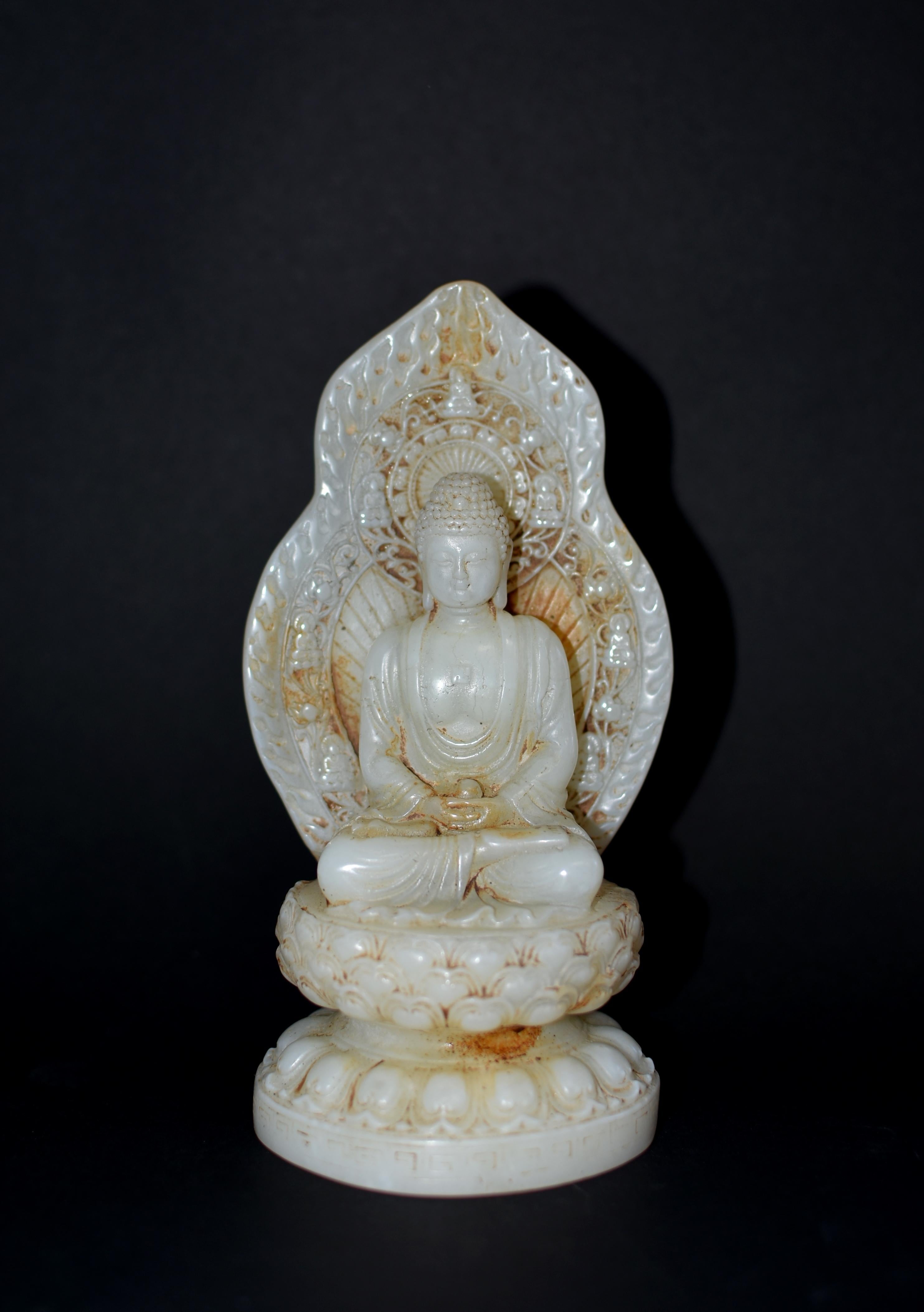 A beautiful statue of white jade Buddha seated in dhyanasana on a lotus base above a waisted round lotiform base. The broad face with smiling downcast eyes casting a serene aura, framed by long-lobed ears and beneath neatly coiled hair. Buddha wears