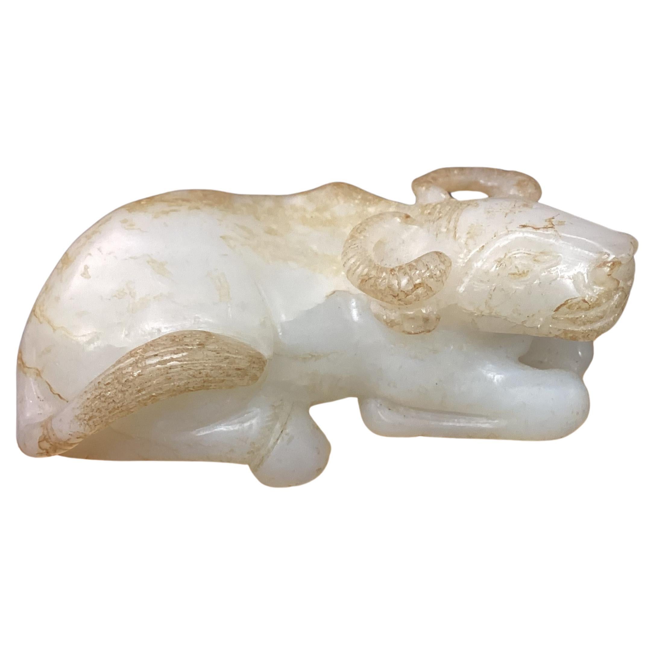 19th century white jade carving of recumbent water buffalo. Buffalo is lying with legs tucked beneath its body, head is raised in an attentive gaze, and its tail is curved over the hind quarters. Color is white with areas of russet and brown
