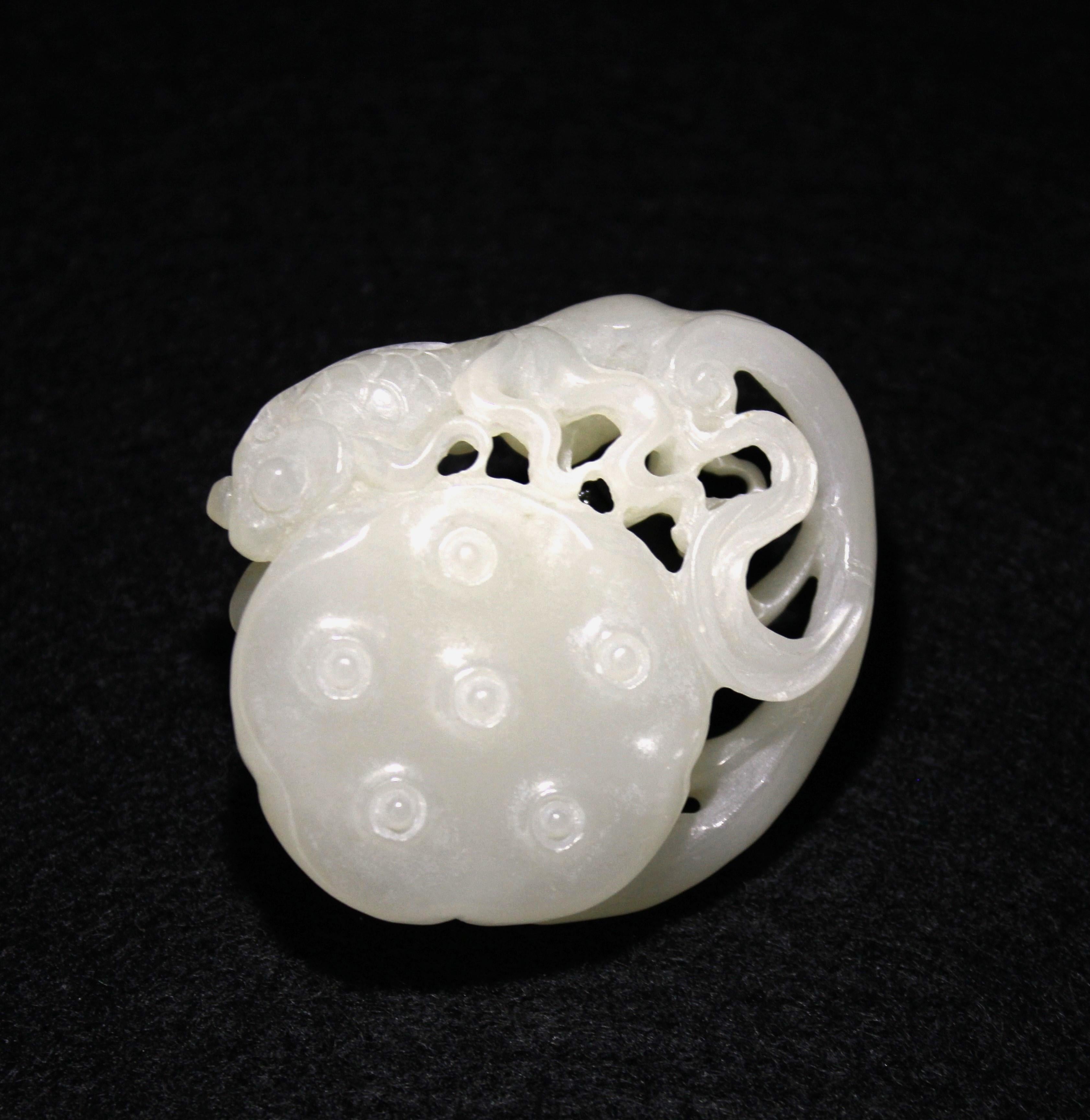 Sculpté dans du jade néphrite, le pendentif représente un bouton de lotus rond sur lequel est gracieusement perché un poisson rouge, dont la longue queue s'enroule élégamment autour du bouton. Le revers présente des champignons lingzhi et des