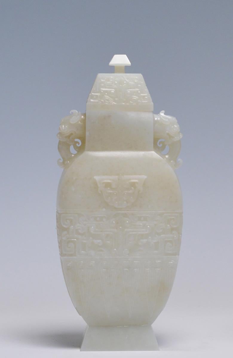 JADE VASE AND COVER 
Of flattened baluster shape, rising from a splayed foot to an angular shoulder and a waisted neck set with a pair of mythical beast heads. Finely carved in low relief around the exterior with a Taotie band, a single mask on each