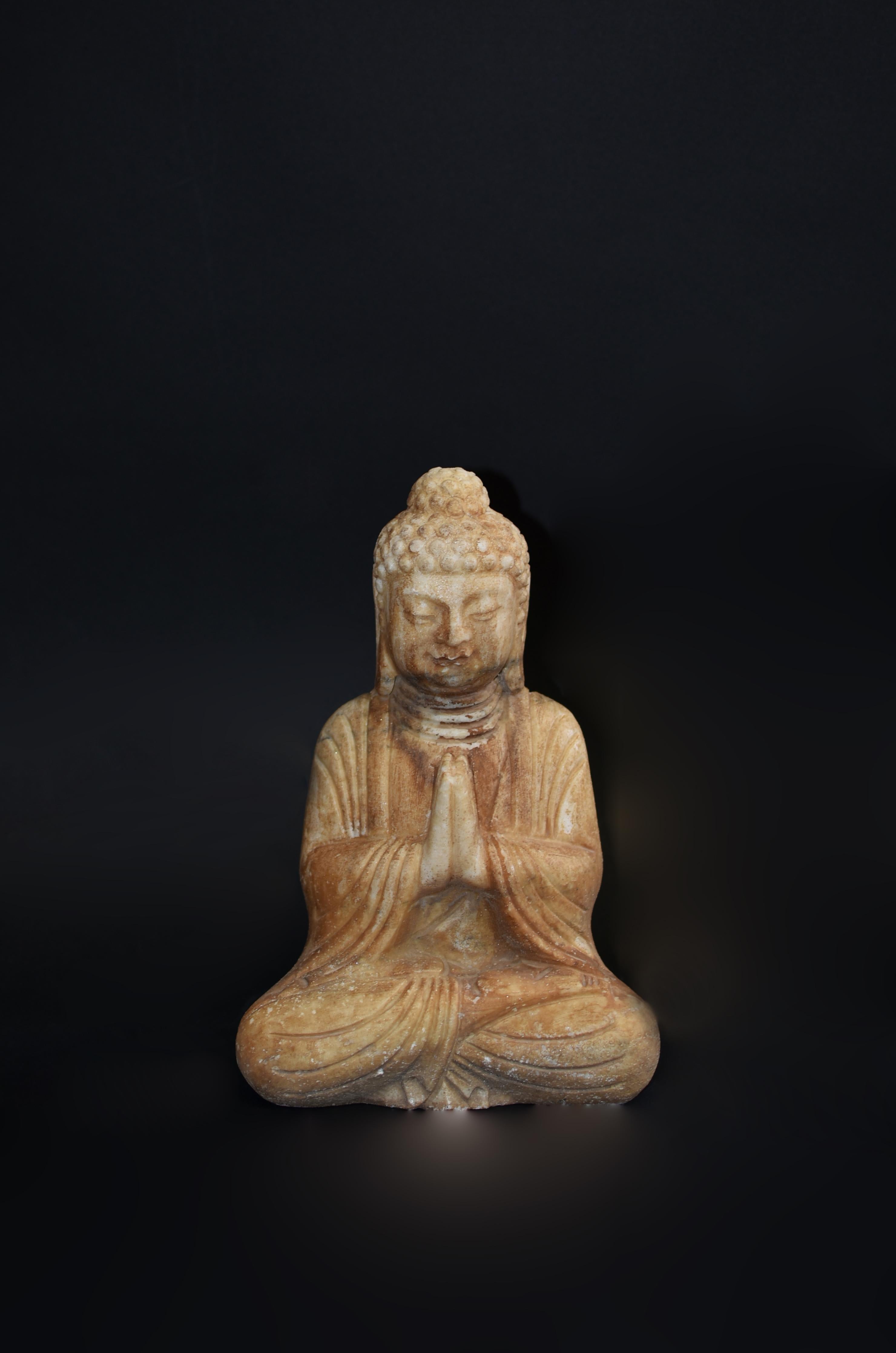 A hand carved solid marble Buddha statue. Seated dhyana asana with hands in namaskar mudra, Buddha has smiling countenance with broad face and large downcast eyes, flanked by pendulous earlobes, conveys sense of peace and harmony. He wears a fluid