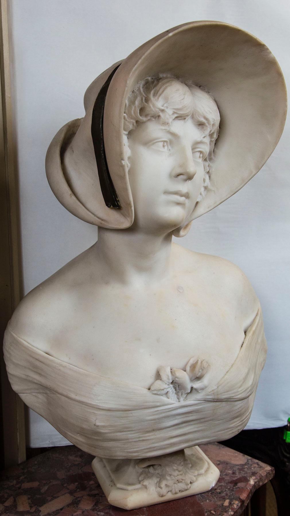 The hat has a bronze band affixed to it. Her shoulders are bare and there is a small bouquet of flowers tucked into the bodice. Her hair cascades from below the back of the hat, which has an upturned brim at the back. Below the bust, on the pedestal