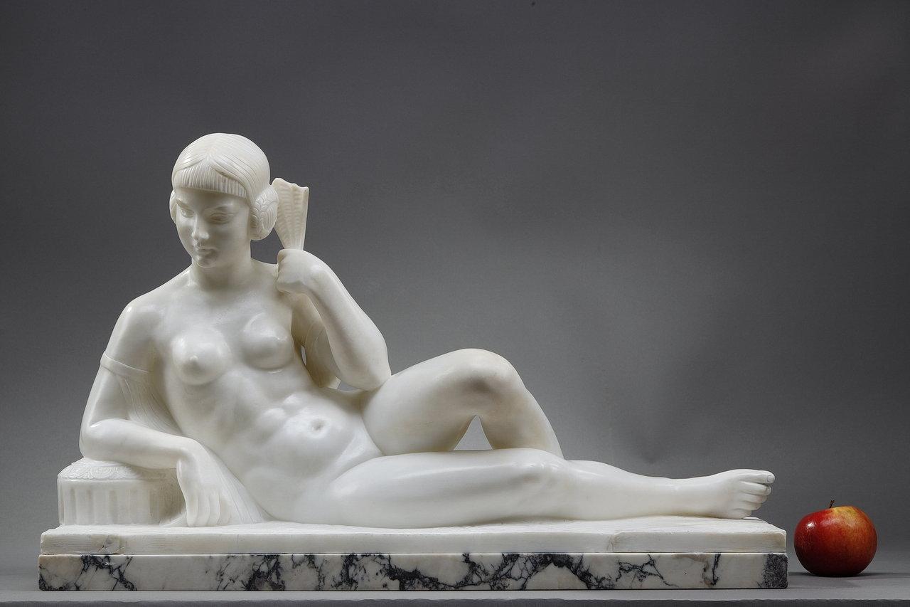 Sculpture Art déco en marbre blanc représentant une odalisque allongée, appuyée sur un coussin et tenant un éventail. La coiffe est stylisée, surmontée d'un bandeau à la mode des années 1930 et maintient des cheveux courts et bouclés. Le travail sur