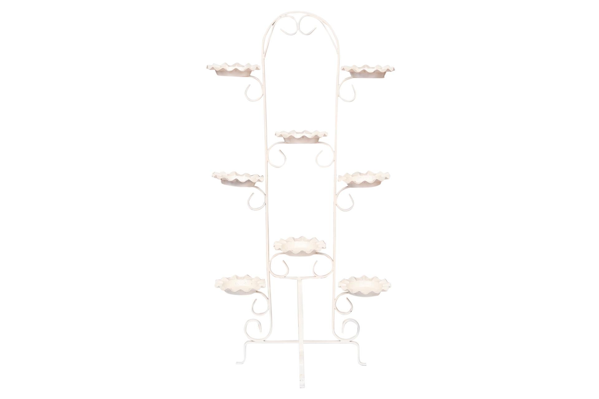 A white metal plant stand. A simple arch supported with scrolls has eight plates for holding plants. The base is formed with two cross pieces shaped into four feet.