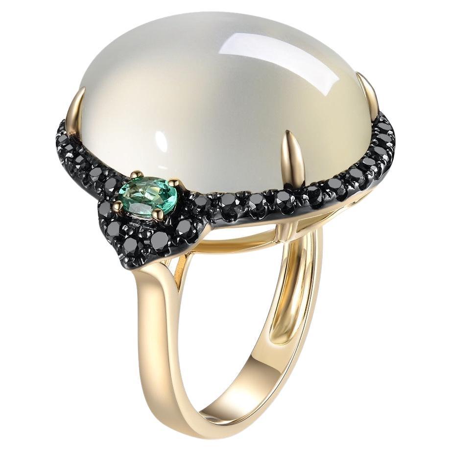 This pretty ring features a large oval moonstone weighing 19.41 carats with beautiful adularescence. . This moonstone is an impressive size, set in a dazzling black diamonds halo flanked with 2 emeralds 14k yellow gold design that showcases its