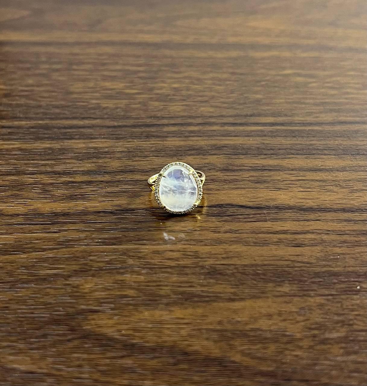 Dieser exquisite Ring zeigt einen fesselnden ovalen weißen Mondstein, eingefasst in ein 14-karätiges Goldband. Der Edelstein ist von einem zarten Halo aus kleinen, funkelnden Diamanten umgeben, die seine Leuchtkraft verstärken. 14,5 X 12 X 4,5 MM