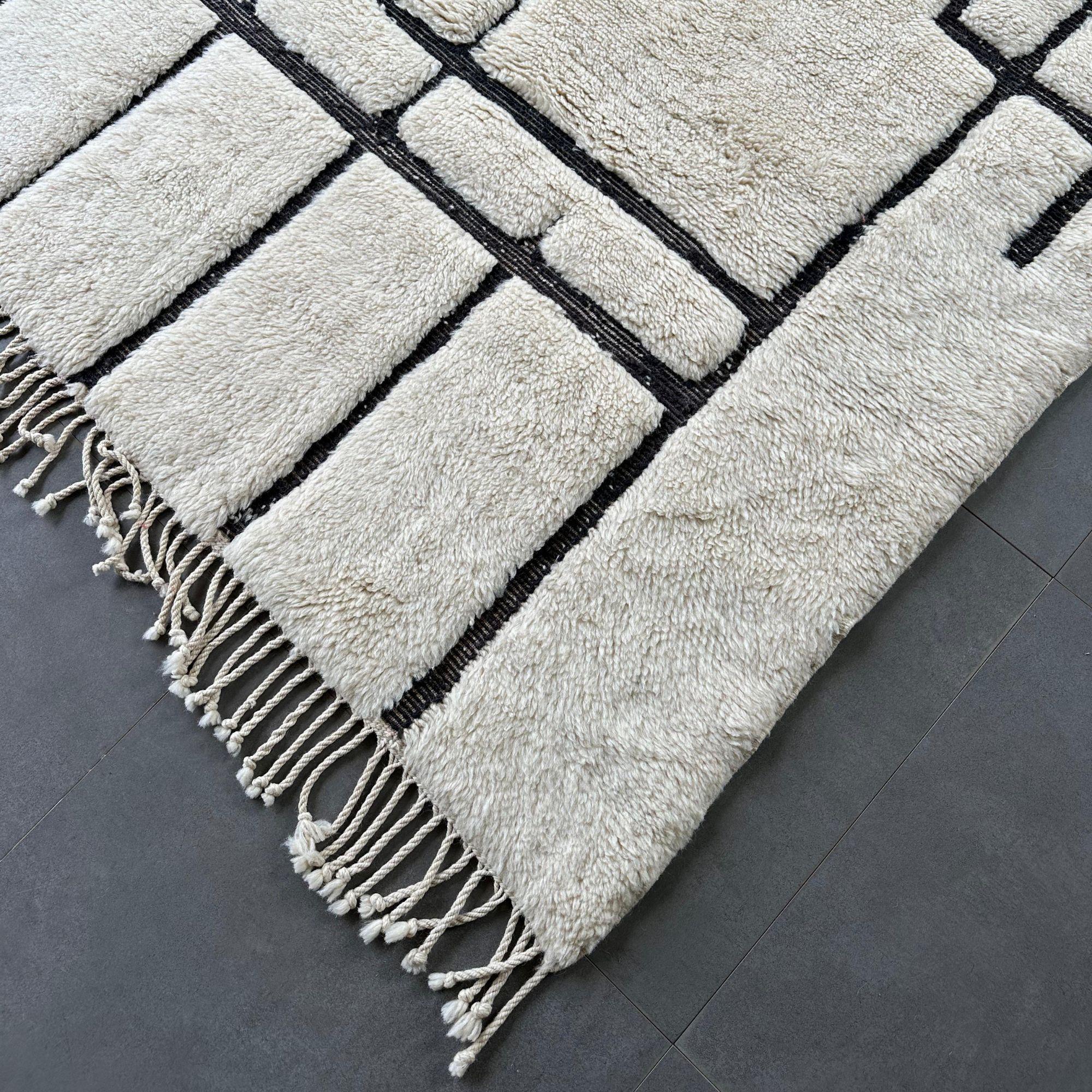 Ti presentiamo Mystic Muse, uno squisito esempio di tappeto marocchino bianco che unisce l'artigianato tradizionale al design moderno. Caratterizzato da una lussuosa base bianca con un audace motivo geometrico nero, questo tappeto aggiunge