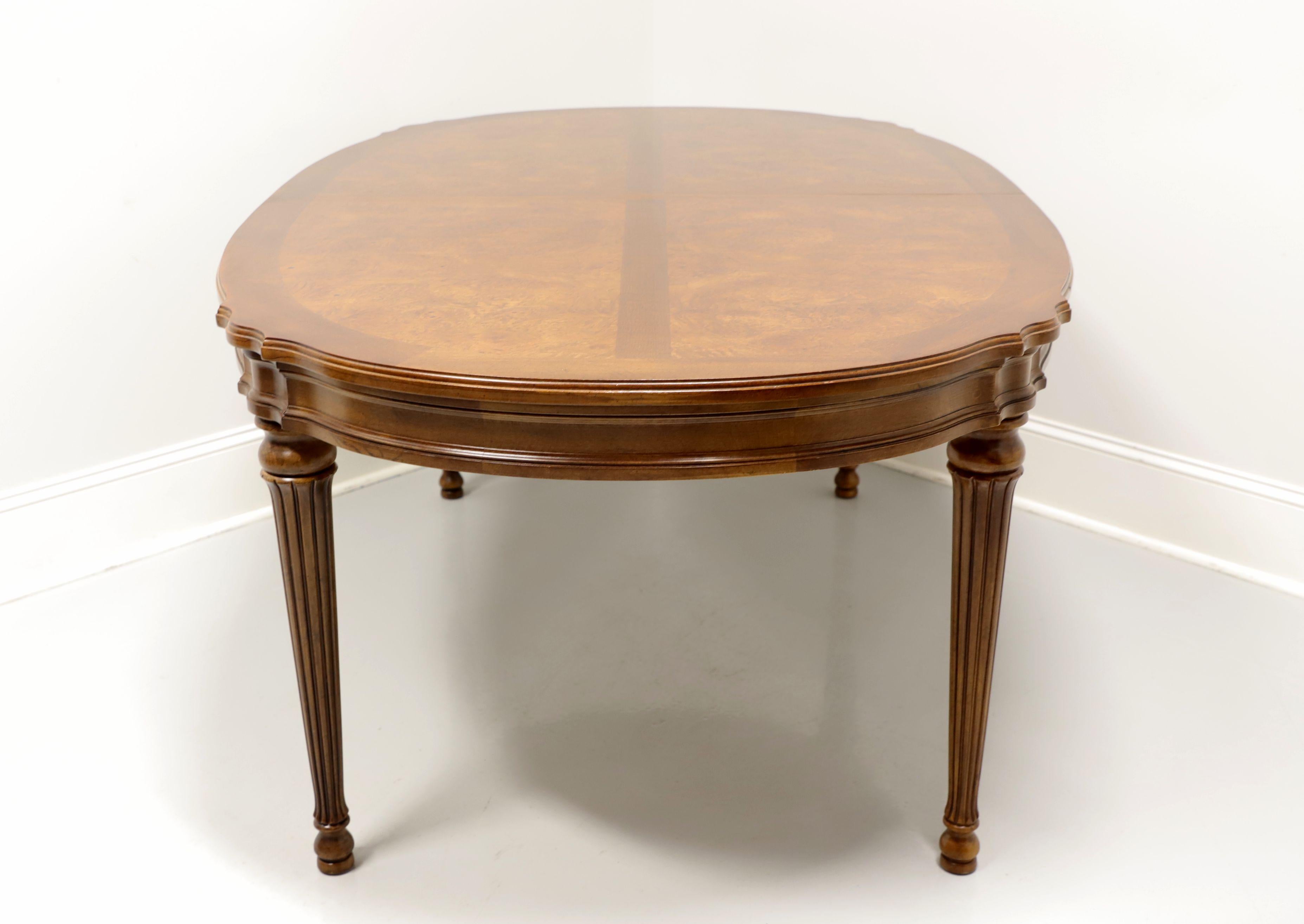 An oval dining table in the French Provincial style by White Furniture Co., of Mebane, North Carolina, USA. Burl Elm with inlaid & banded top, serpentine edge, carved apron, fluted tapered legs and pad feet. Metal expansion sliders. Includes two