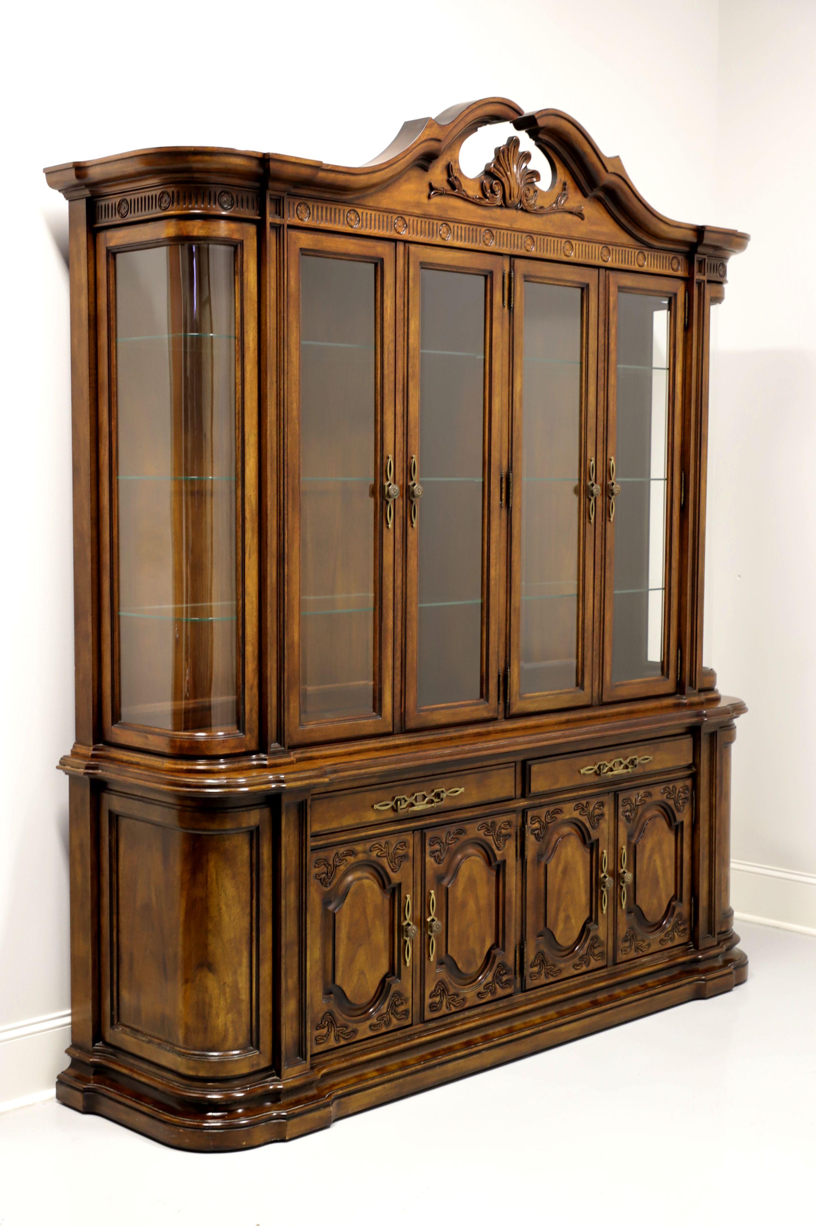 A china display cabinet in the French Country style by White Furniture. Walnut and burl walnut with brass hardware, decoratively carved pediment top, carved frieze at top, stepped ogee edge at middle between cabinets, rounded corners, and a solid