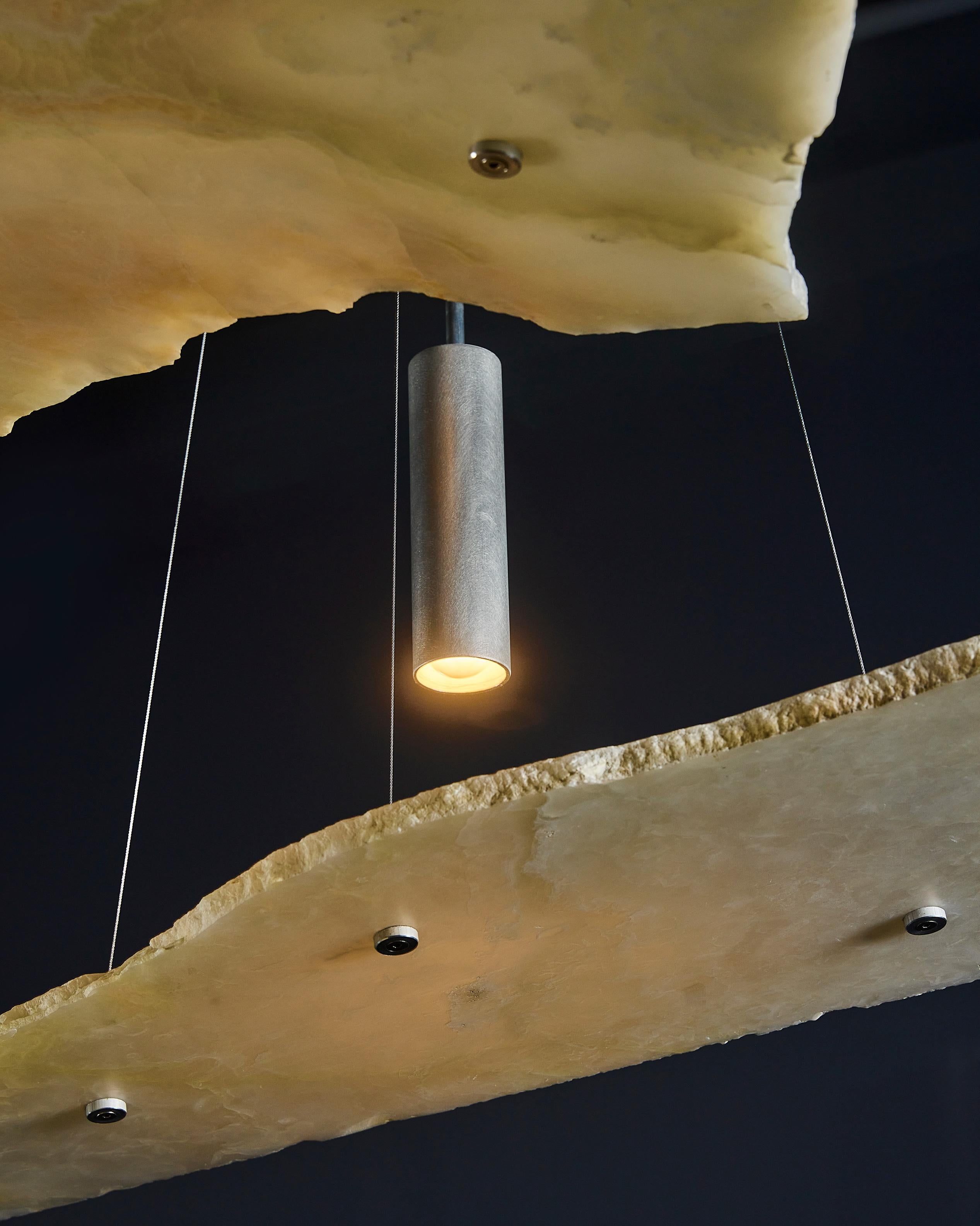 Le lustre Unbroken utilise de l'onyx blanc à bords bruts en suspension pour créer une expérience visuelle puissante avec la lumière. Les arêtes naturelles de la pierre sont laissées apparentes et contrastent avec la surface adoucie de la dalle. En