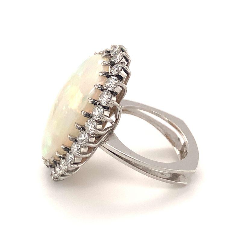 This bold and glowing 1970s white opal and diamond ring is an exquisite statement piece, set in luxurious 18K white gold. At its center lies a magnificent 14-carat oval cabochon opal, radiating a soft, ethereal glow with flashes of iridescent color