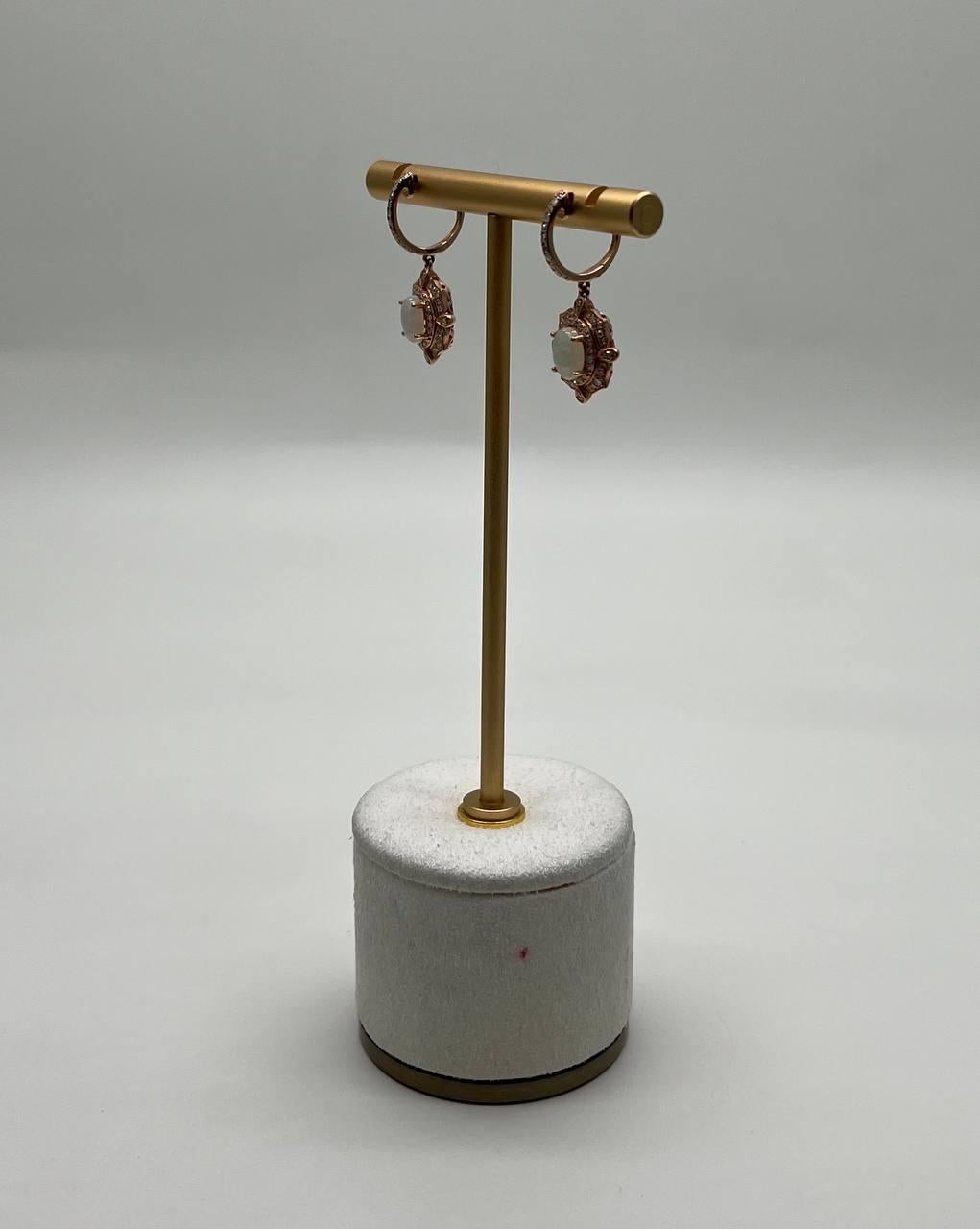 These sophisticated earrings are crafted with precision and artistry.
Each earring features a central oval white opal, set within a detailed, ornate frame.
The frame is adorned with small, sparkling diamonds that enhance the central stone's