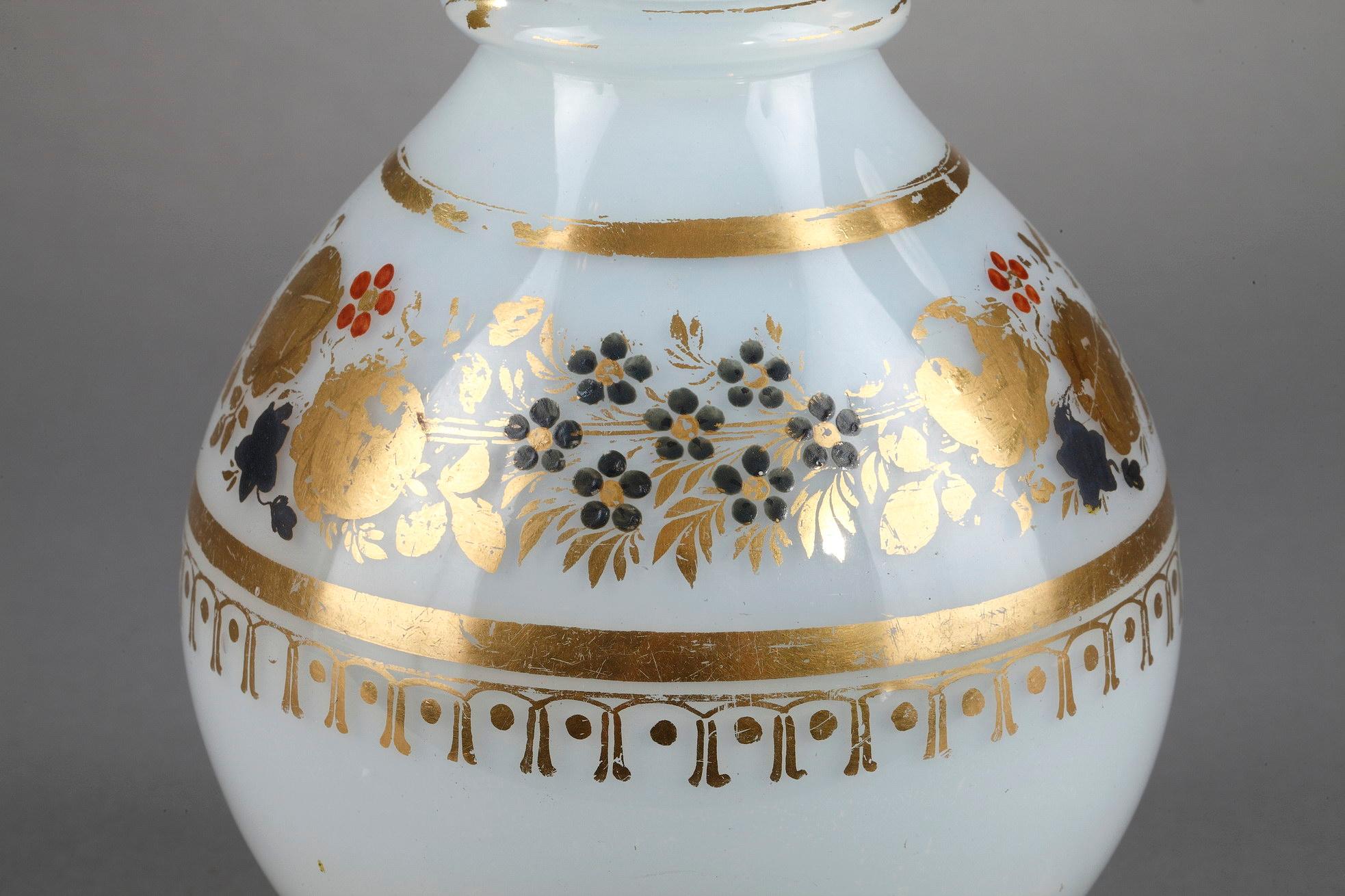 White opaline bottle with a ball-shaped stopper. Golden stripes, gold, and a dark blue wreath of roses, anemones, and forget-me-nots decorate the rim of the paunch. The top of the stopper is highlighted with petals. he delicate decoration was