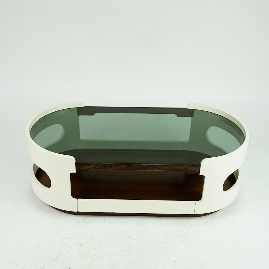 This small fantastic oval Space Age coffee or cocktail table has been designed and produced in Italy in the 1960s. It features a oval white lacquered plywood base with a glass top and rosewood foot and shelve.
It´s style is very close to Joe