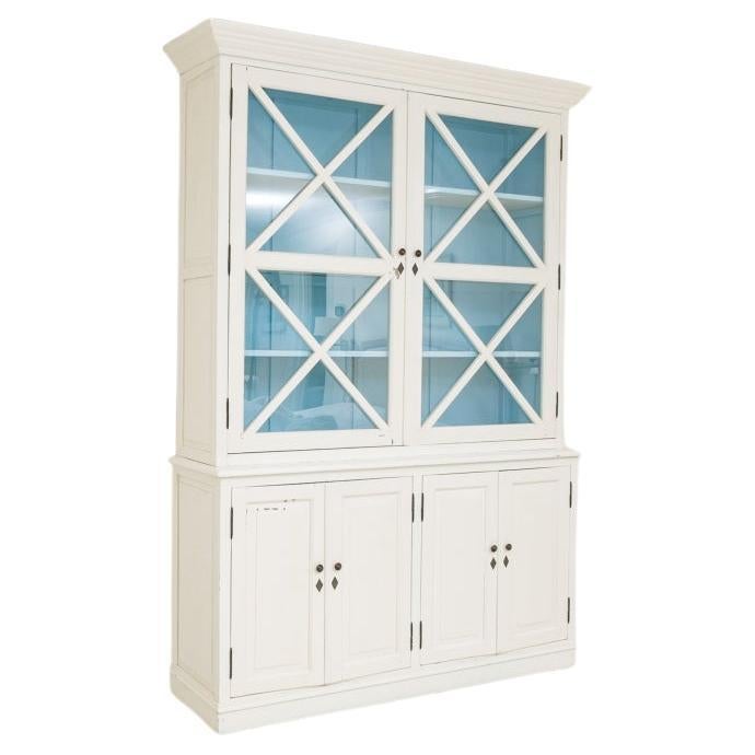 Armario de época decorado con pintura blanca y dividido en dos partes; el armario superior tiene dos puertas acristaladas con parteluces en forma de X y molduras de corona. Las puertas acristaladas se abren a un interior entarimado pintado de vivo