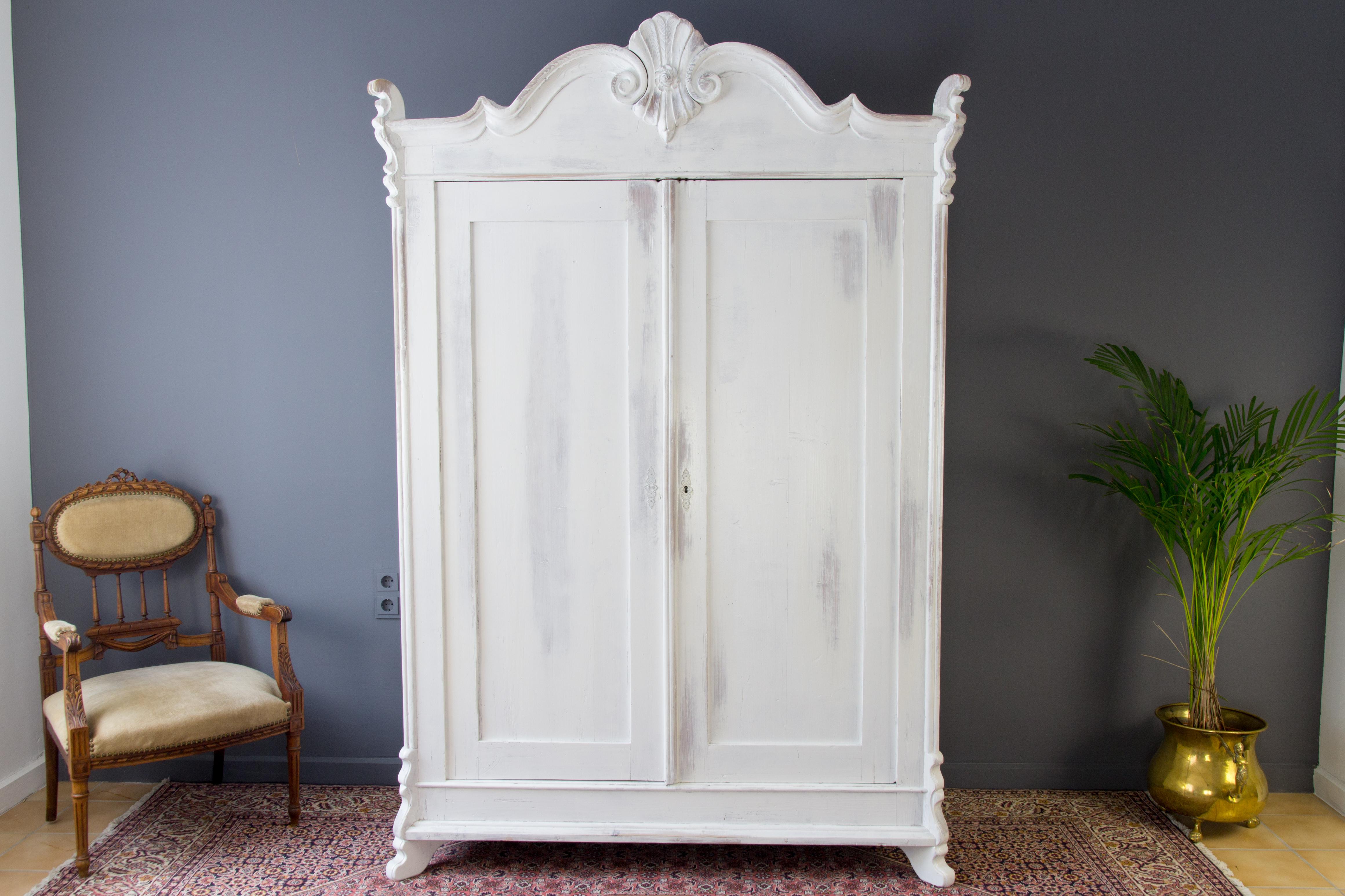 Two-door armoire of Baltic pine with five shelves. The exterior front side is white painted. Inside, the pine remains in its raw color. One side is equipped with five shelves, the other - with original clothes hangers and one shelf. The wardrobe is