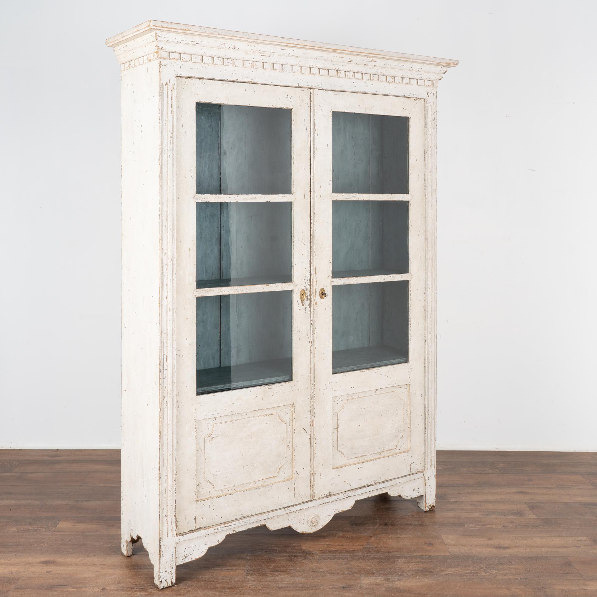 Delightful white painted pine bookcase with dentil molding along crown and fluted carved details along flanking sides. 
The simple lower panels and carved skirt add balance to this display cabinet.
Restored, later professionally painted in layered