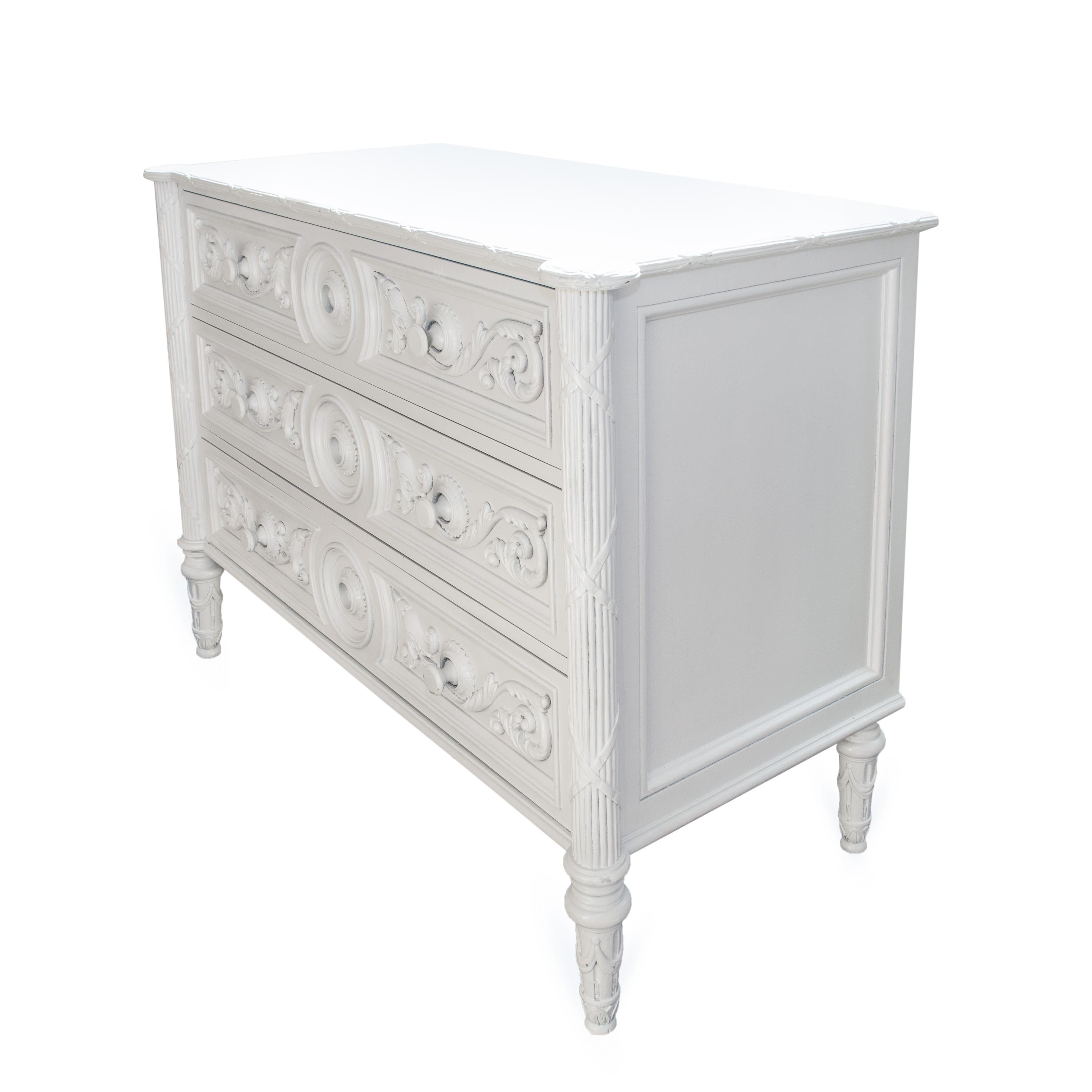 This is a white-painted American chest of drawers. The top has a rounded edge over three long graduated drawers with carved ornamentation on the drawer faces and supported by round tapered feet. The sides are paneled, 20th century.