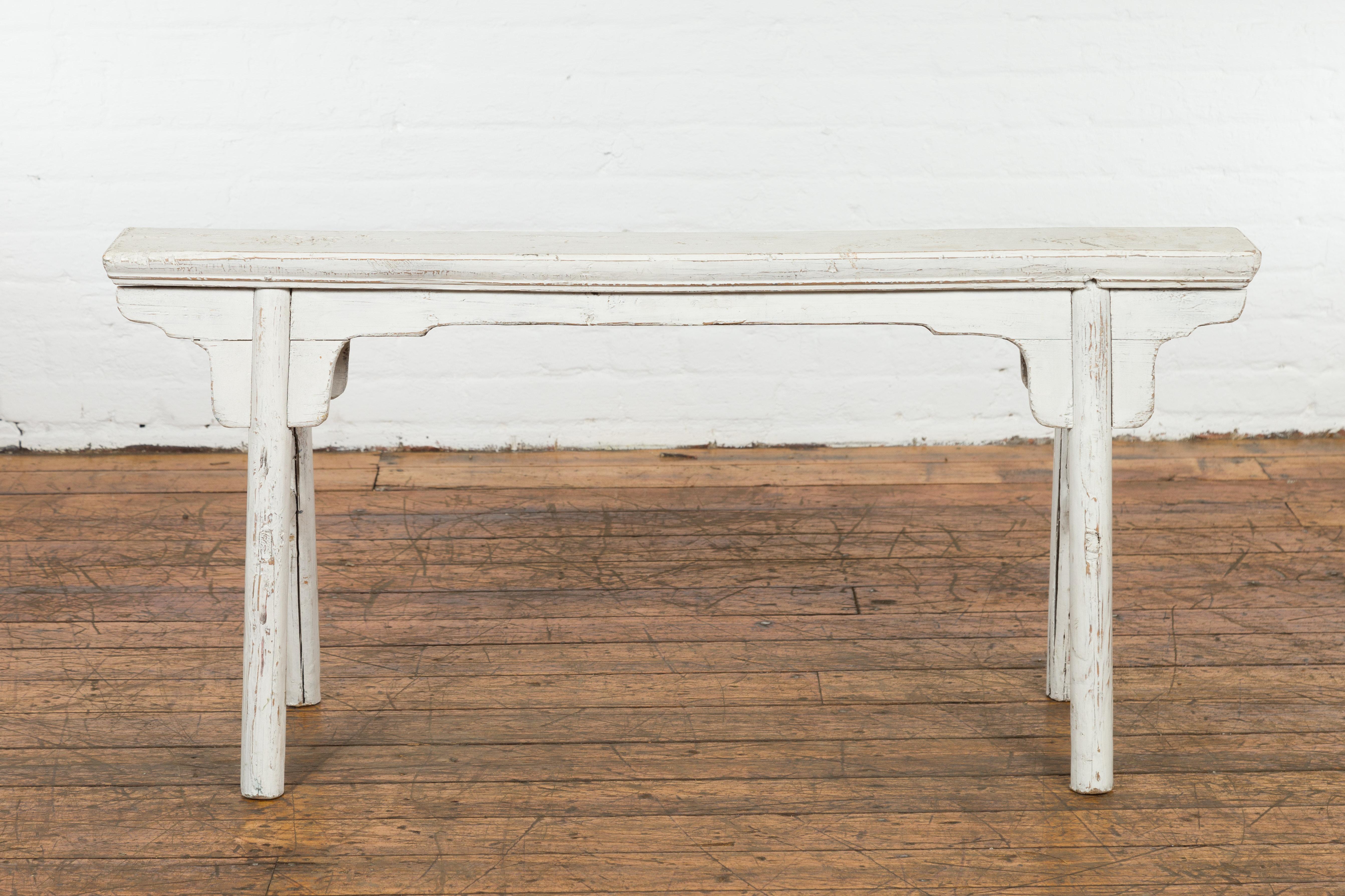 A Chinese vintage Ming Dynasty style white painted wooden bench from the mid 20th century with A-form base, carved spandrels and distressed finish. Created in China during the Midcentury period, this small bench draws the attention with its soft