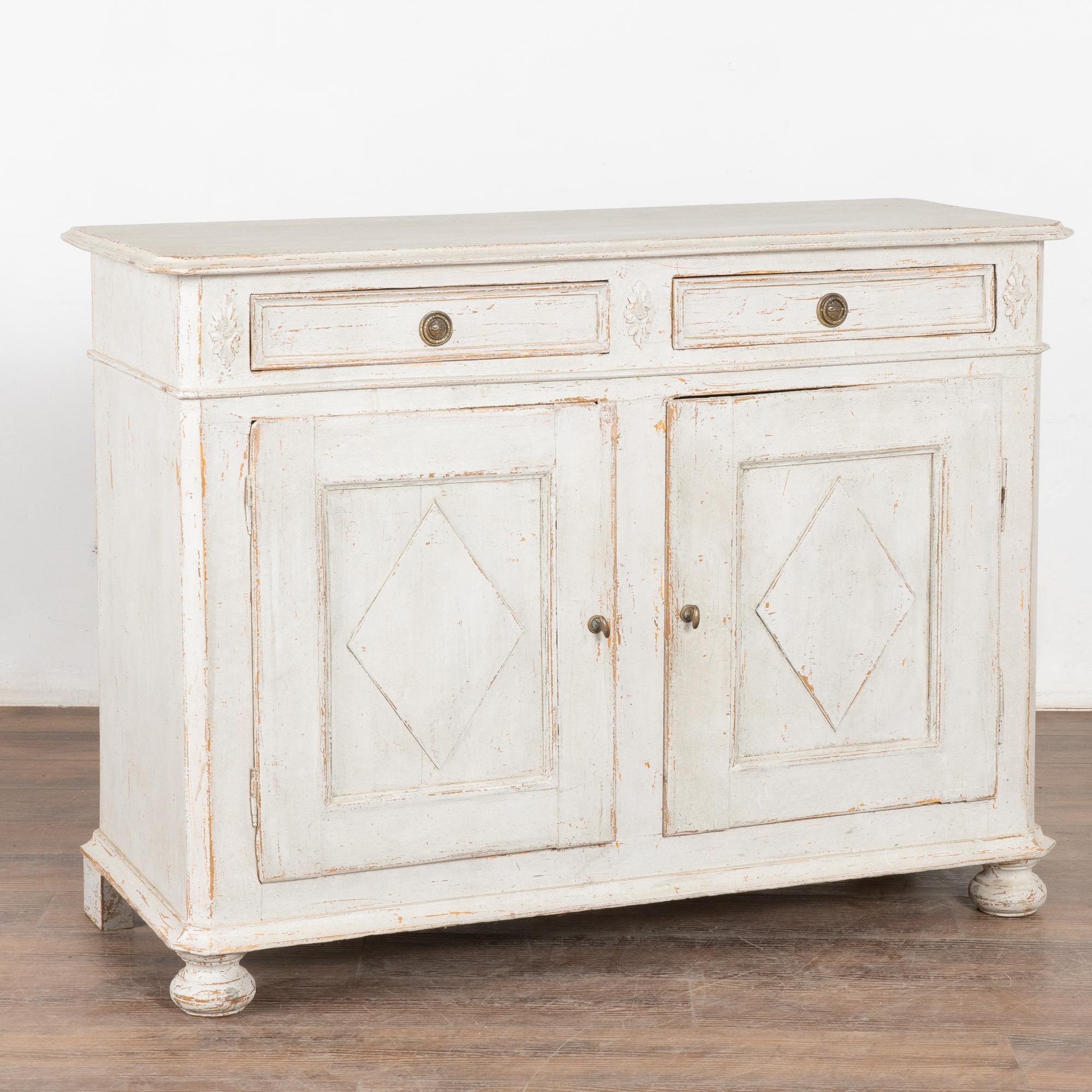 Gustavian pine sideboard or buffet with traditional decorative diamond panels on cabinet doors. 
Later professionally painted in layered shades of antique white and lightly distressed to fit the age and grace of this lovely sideboard.
Brass pulls on