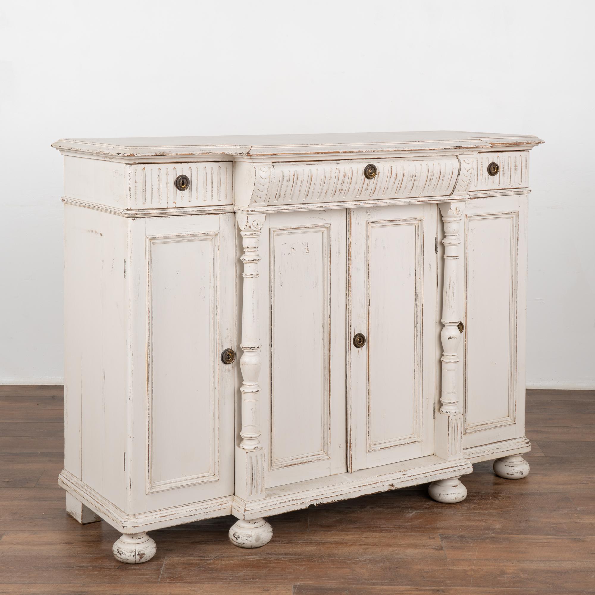 Swedish pine sideboard with turned columns, top with profiled edge, fluted carving along drawers all resting on large bun feet.
Newer, professionally applied white painted finish slightly distressed to fit the age and grace of this lovely