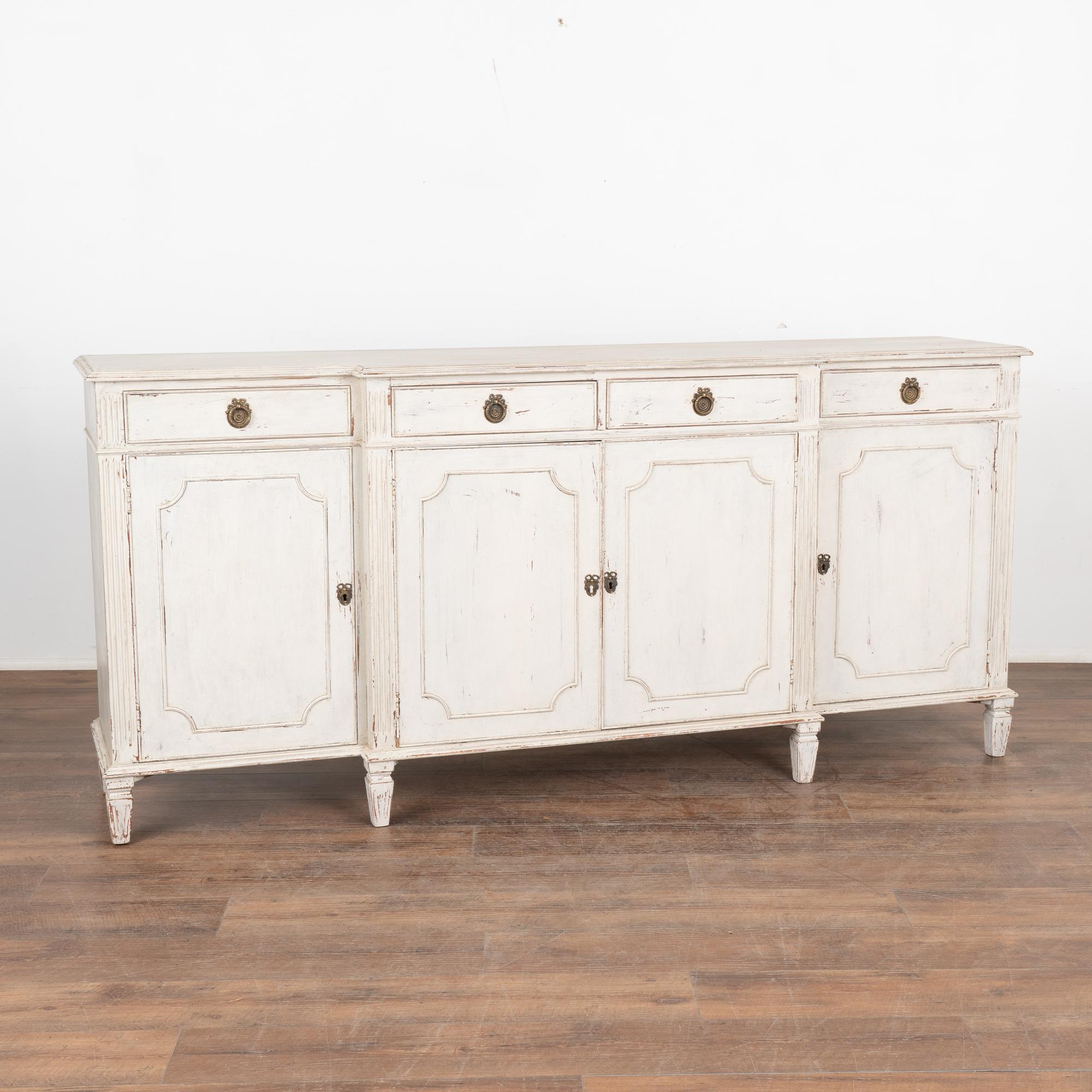 Swedish white painted sideboard/buffet with paneled doors and fluted tapered feet.
Four drawers with brass pulls over four cabinet doors, one interior shelf.
Restored, later professionally painted in layered shades of antique white and lightly
