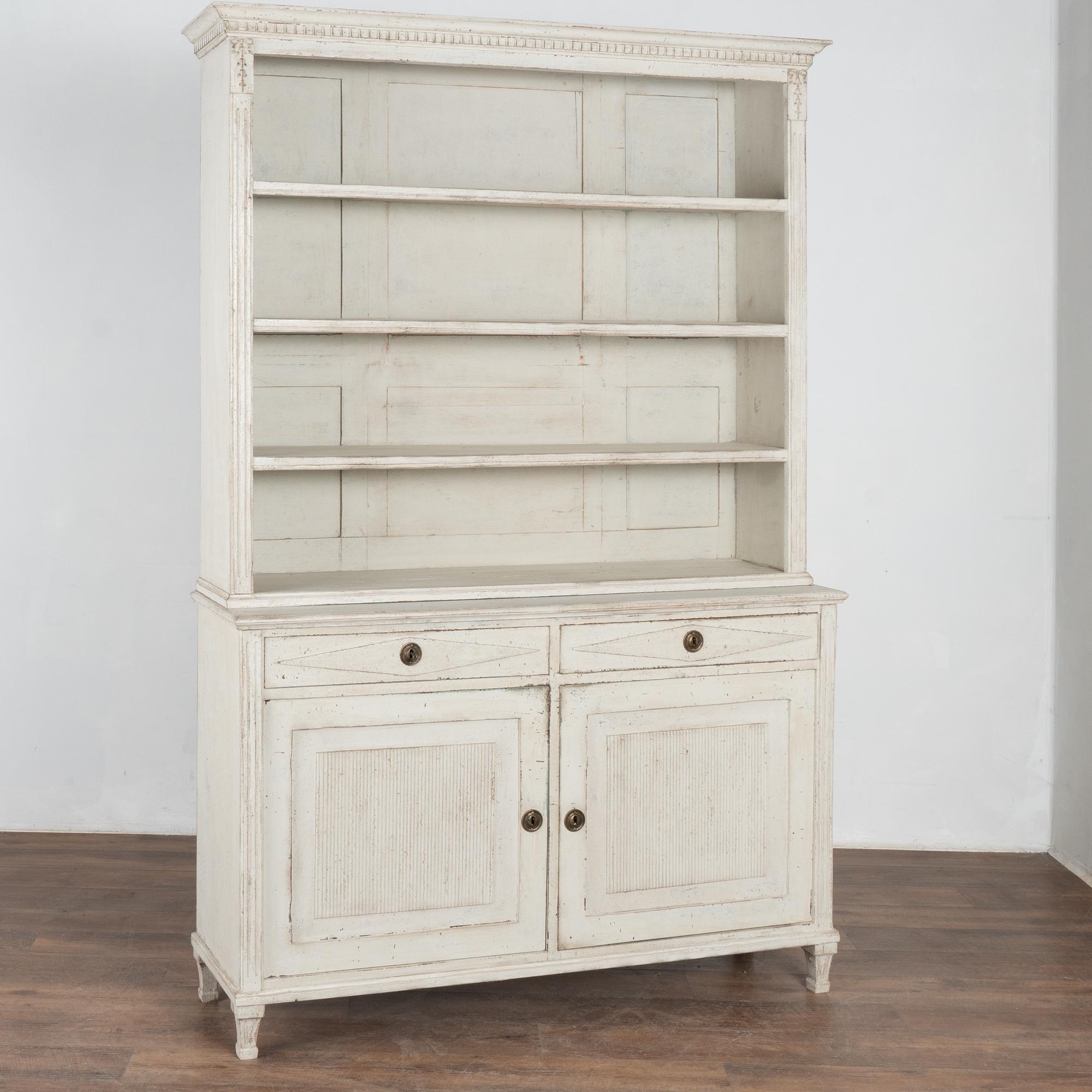 This pine cupboard may be used as a display cabinet or bookcase. Dentil molding, diamonds on the drawers and fluted carving along doors and sides reflect its Swedish country styling.
Restored, later professionally painted in layered shades of white