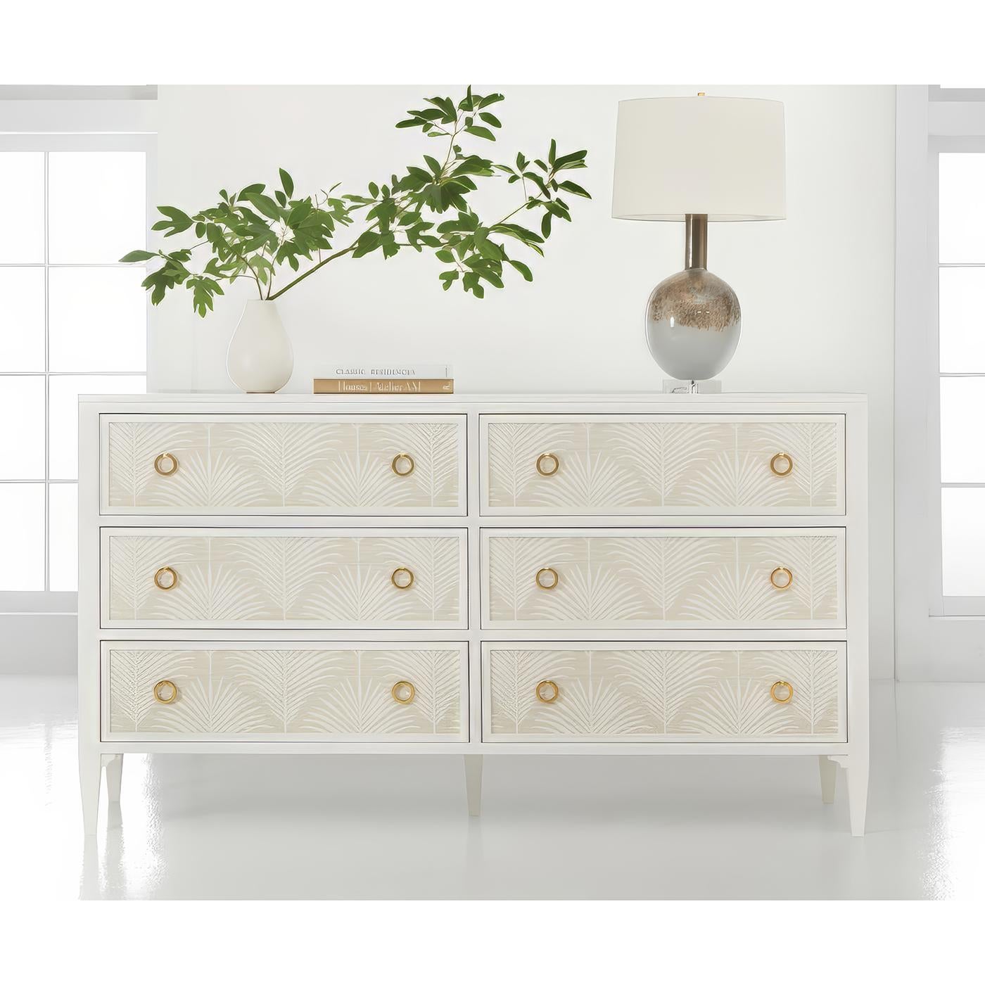 Presenta intricati motivi a fronde di palma applicati ai frontali dei cassetti. Il design sofisticato abbina il legno verniciato di bianco con i tiranti ad anello in ottone, mentre le gambe affusolate e le linee pulite creano una silhouette
