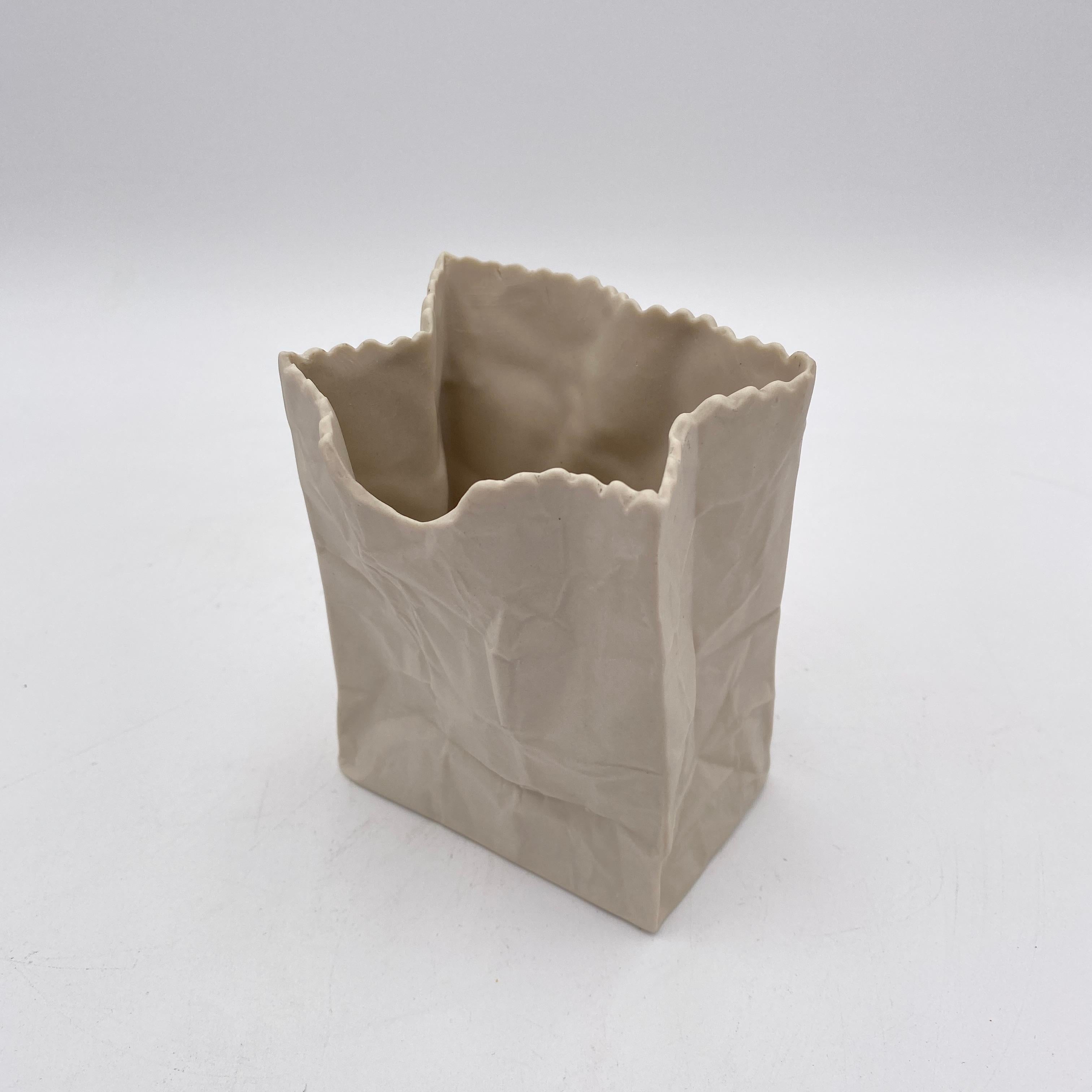Se trata de un diminuto jarrón de cerámica blanco para bolsas de papel. Lleva estampado en el interior, Ceramic Illusions, y se fabricó en California en la década de 1970. Es del estilo del jarrón bolsa de papel de Tapio Wirkkala para Rosenthal.