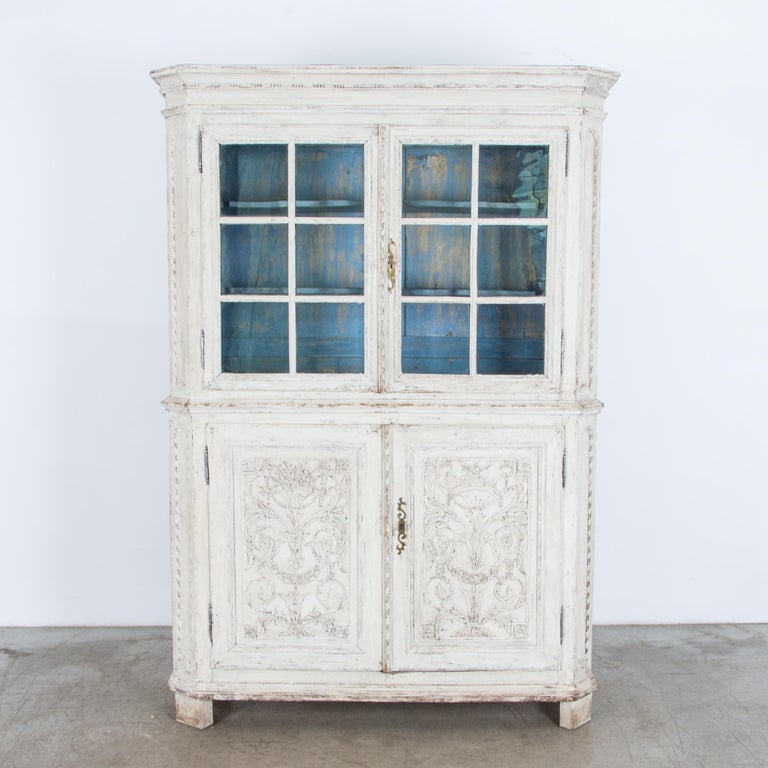 Two lower doors and glass display windows give this piece potential for storage or display. From France circa 1800, an ornately carved decorative motive becomes a dramatic ground for rustic painting, in glossy antique white gently distressed.