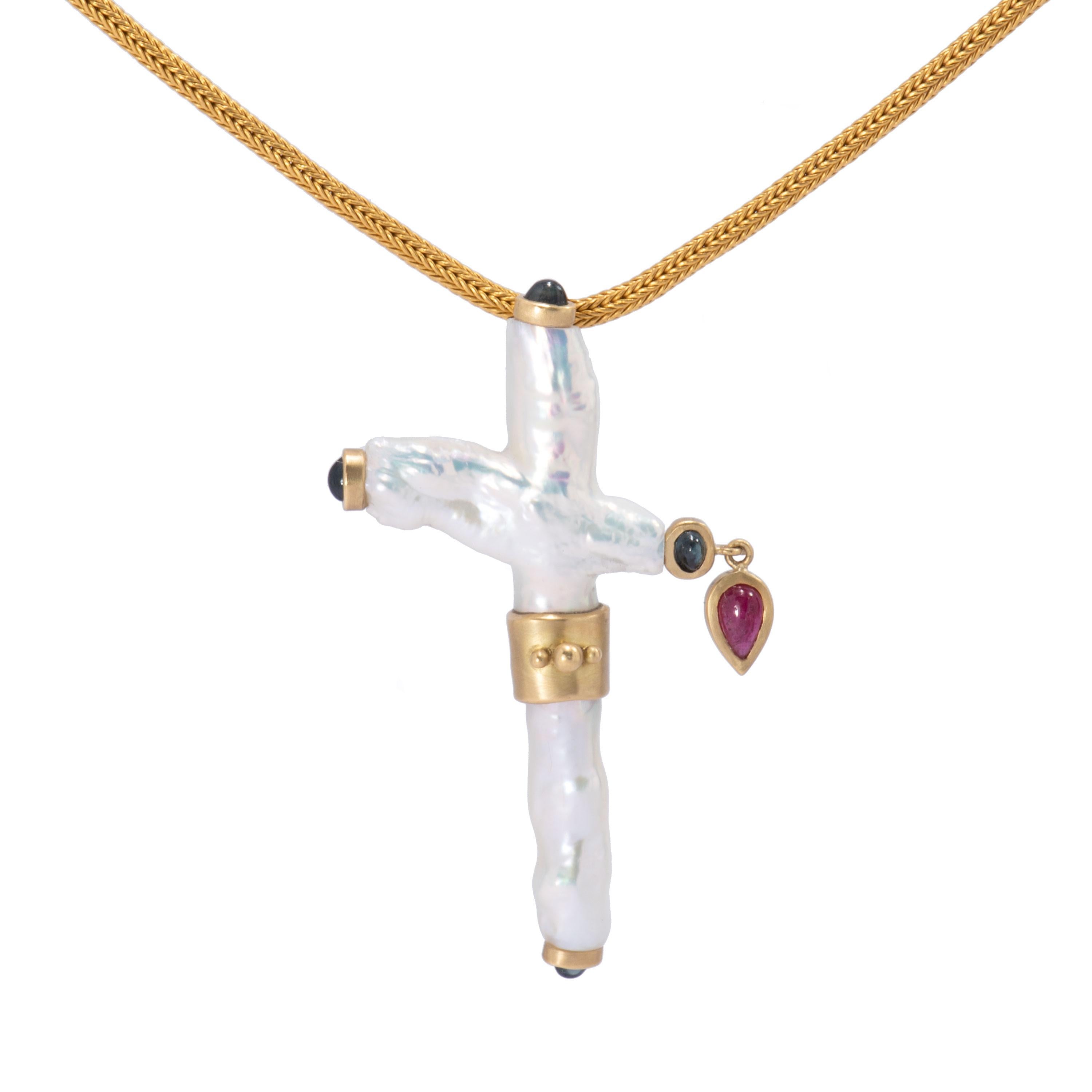 A naturally formed white pearl in the shape of a cross is bezel set in 18 karat gold with 4 deep blue sapphire cabochons at the 4 stations. A tear shaped ruby cabochon dangles from one station and an 18 karat gold beaded band encircles the center.