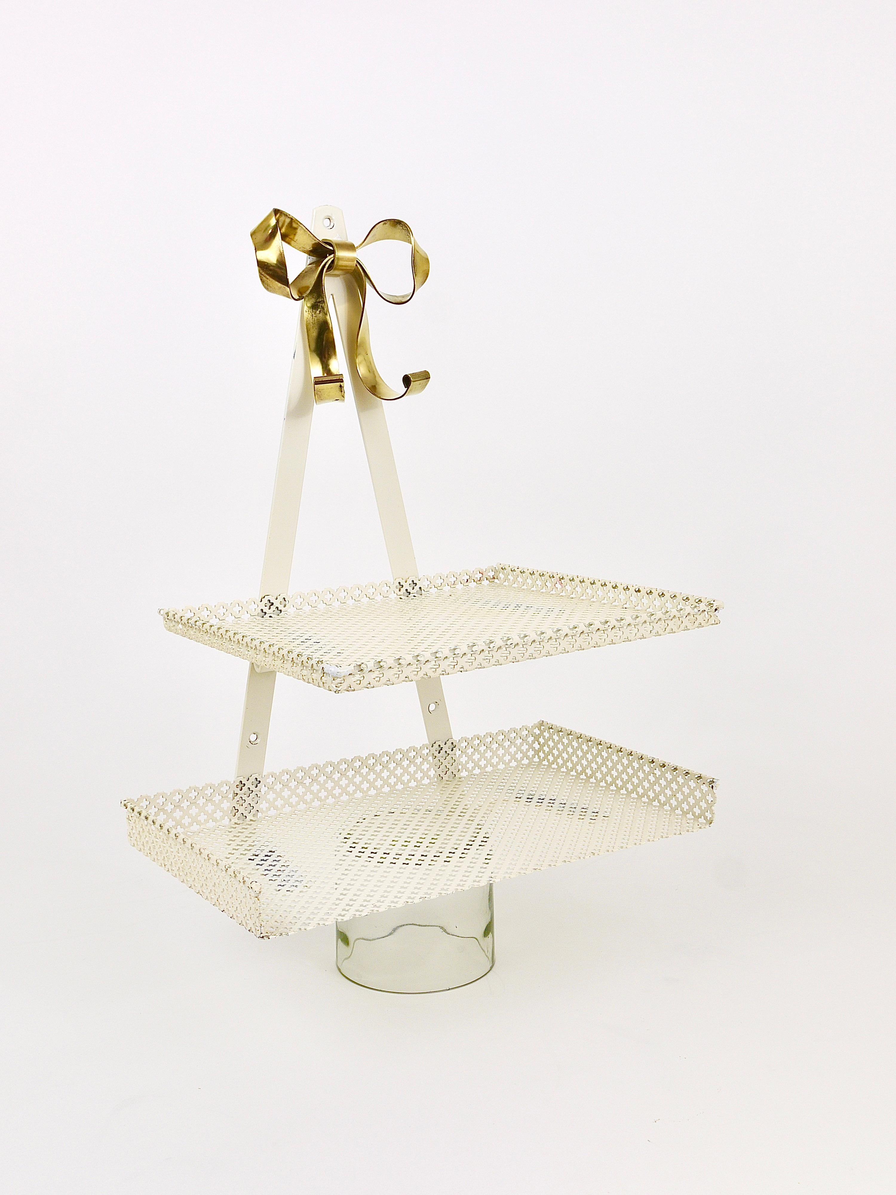 A beautiful wall-mounted midcentury rack with two shelves and a lovely brass ribbon on its top. Manufactured in the 1950s by Vereinigte Werkstätten München / Munich / Münchner Werkstätten, Germany. Made of white lacquered perforated metal and