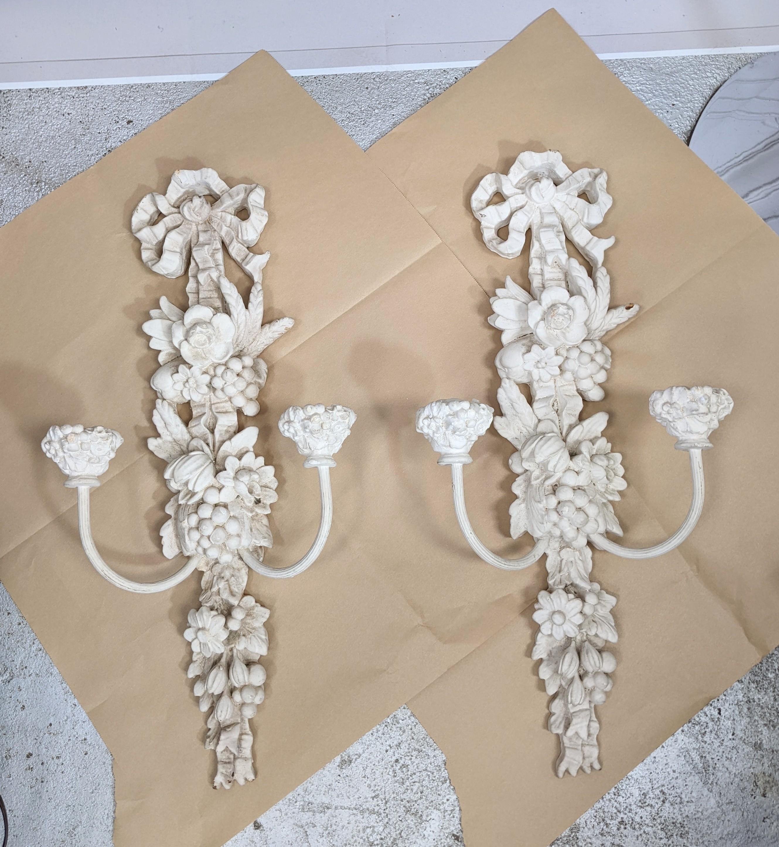 Pair of Hollywood Regency style plaster on wood wall candle sconces from the 1940's. Fully formed, dimensional motifs of fruit and flowers hang from ribbon bow motifs. Plaster molded on wood backing with enamel metal arms. 1940's USA.