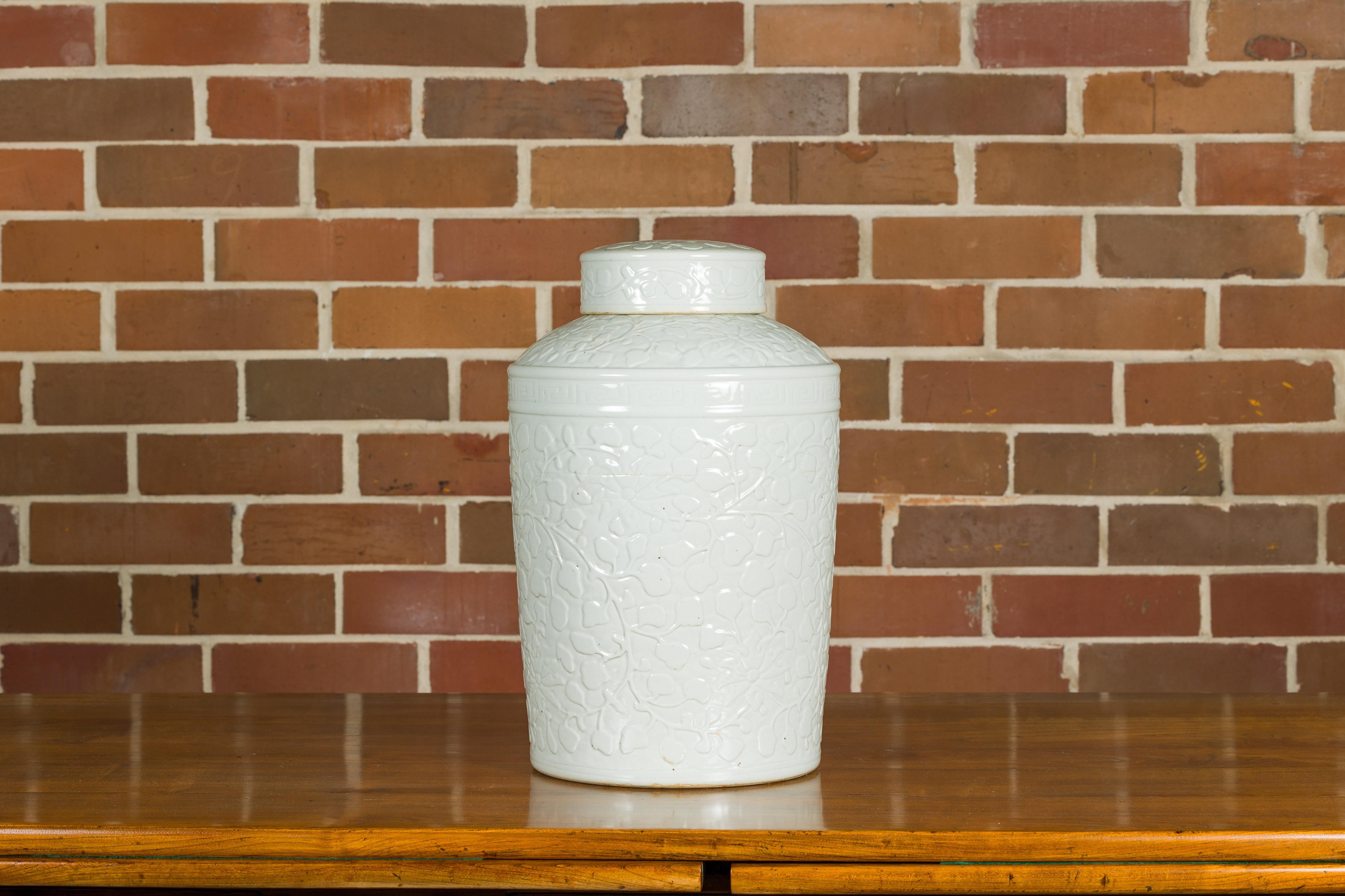 A white porcelain Asian lidded jar with scrolling foliage motifs and slightly tapering lines. Behold the serene elegance of this white porcelain Asian lidded jar, a piece that gracefully marries simplicity with ornate detailing. Adorned with
