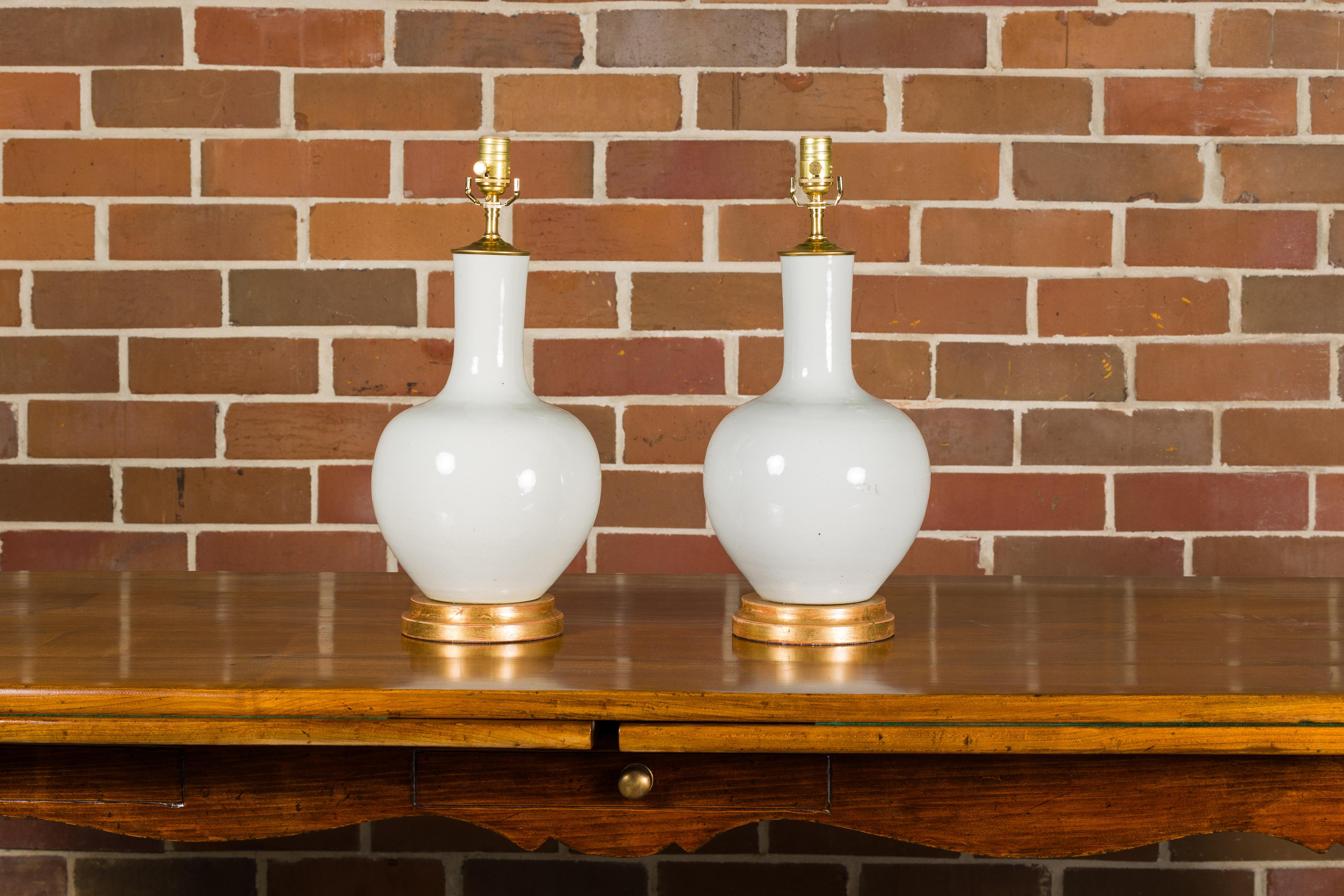A pair of white porcelain vases made into table lamps wired for the USA and mounted on giltwood circular bases. Elevate your living space with this pair of white porcelain vase table lamps, skillfully adapted for use in the USA and exquisitely
