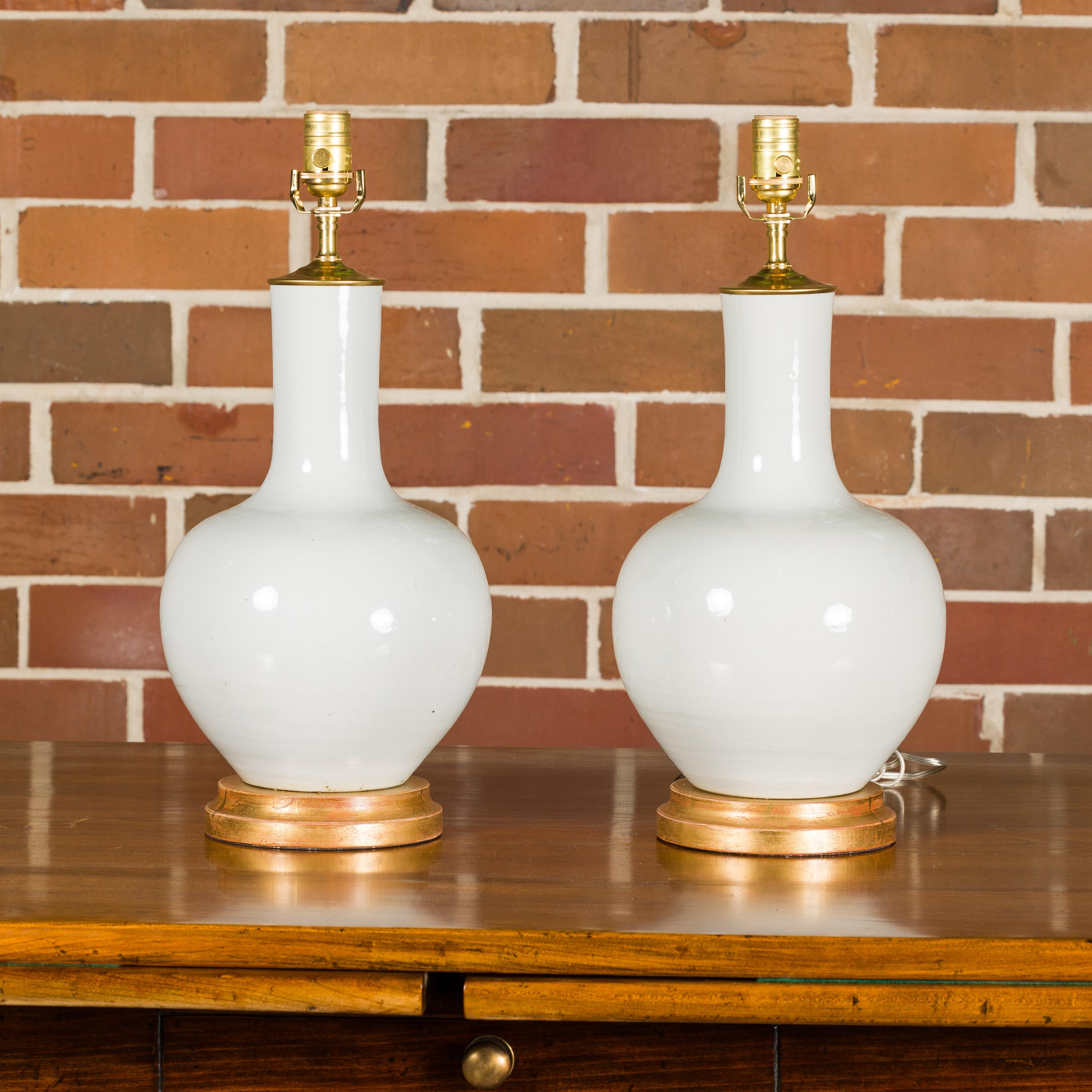 A pair of white porcelain vases made into table lamps wired for the USA and mounted on giltwood circular bases. This pair of table lamps seamlessly blends classic elegance with modern functionality. Crafted from pristine white porcelain, these vases