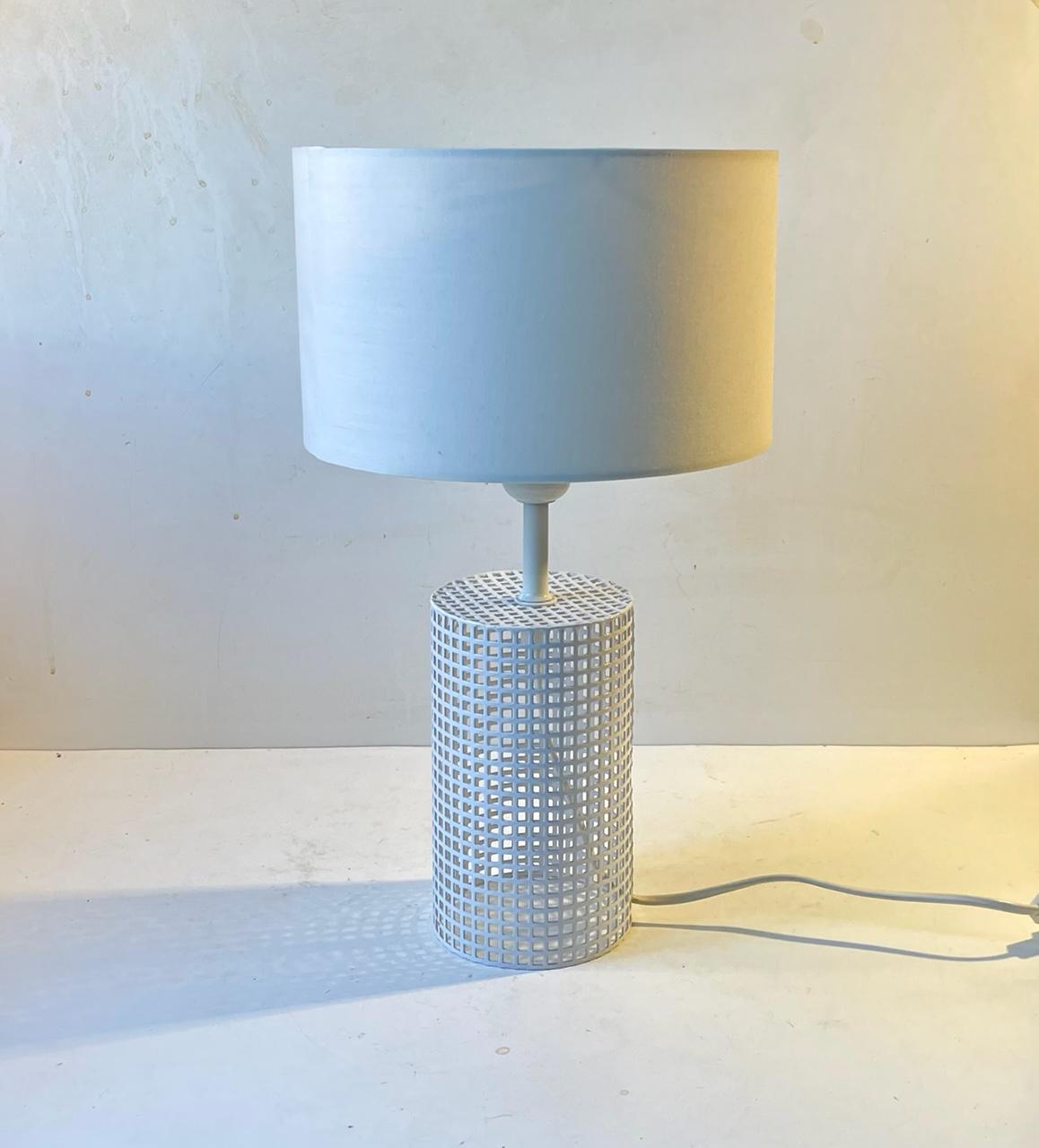 A stylish and clean white table lamp featuring a pierced/perforated metal base set white a cylindrical white textile shade. It has a simple on/of switch to its cord. Designed and manufactured by Laoni Belysning in Denmark during the 1970s in a style