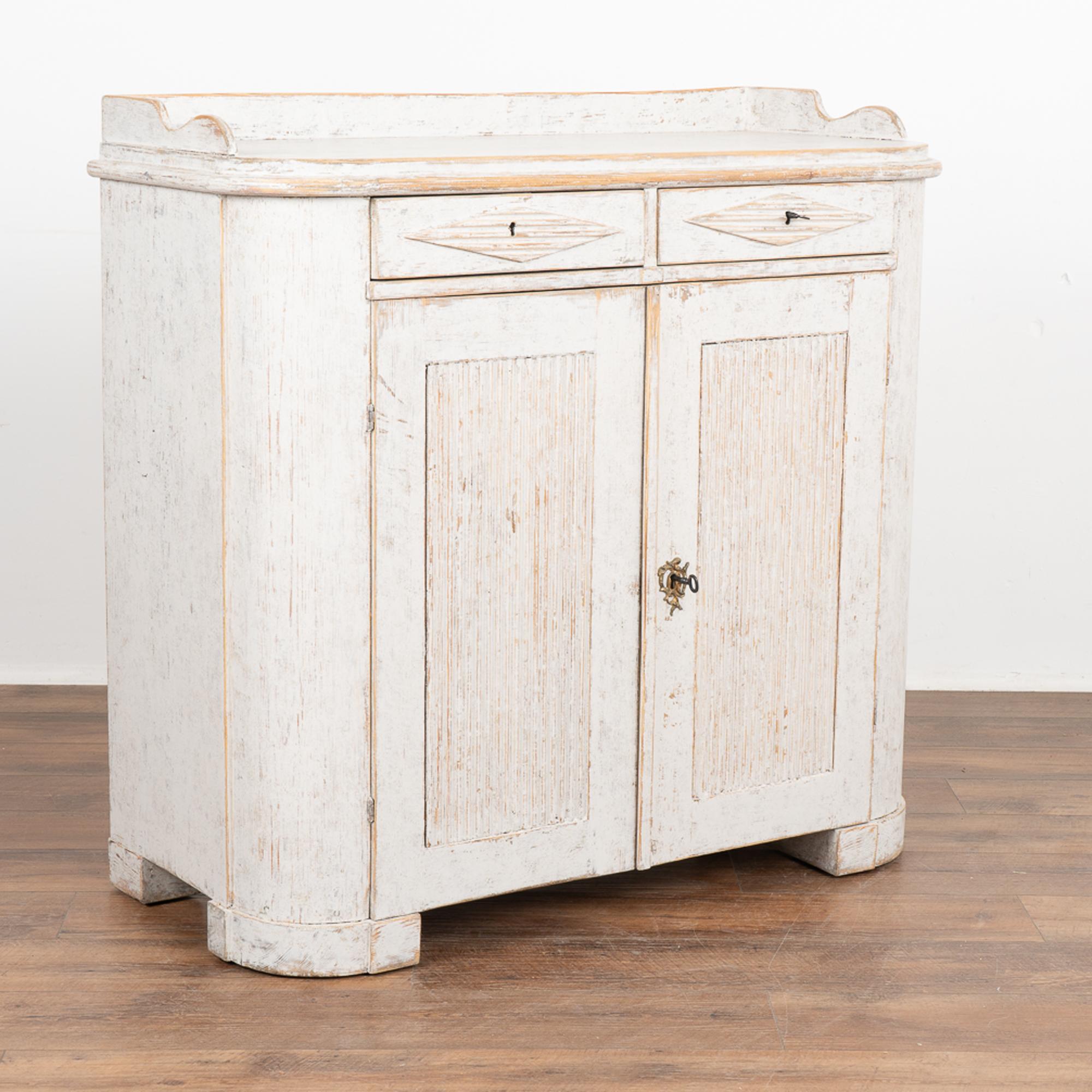 Gustavian small pine sideboard with traditional decorative fluted panels on cabinet doors, two small upper drawers with fluted diamond detail.
The newer, professionally applied white exterior is slightly distressed, fitting the age and grace of this