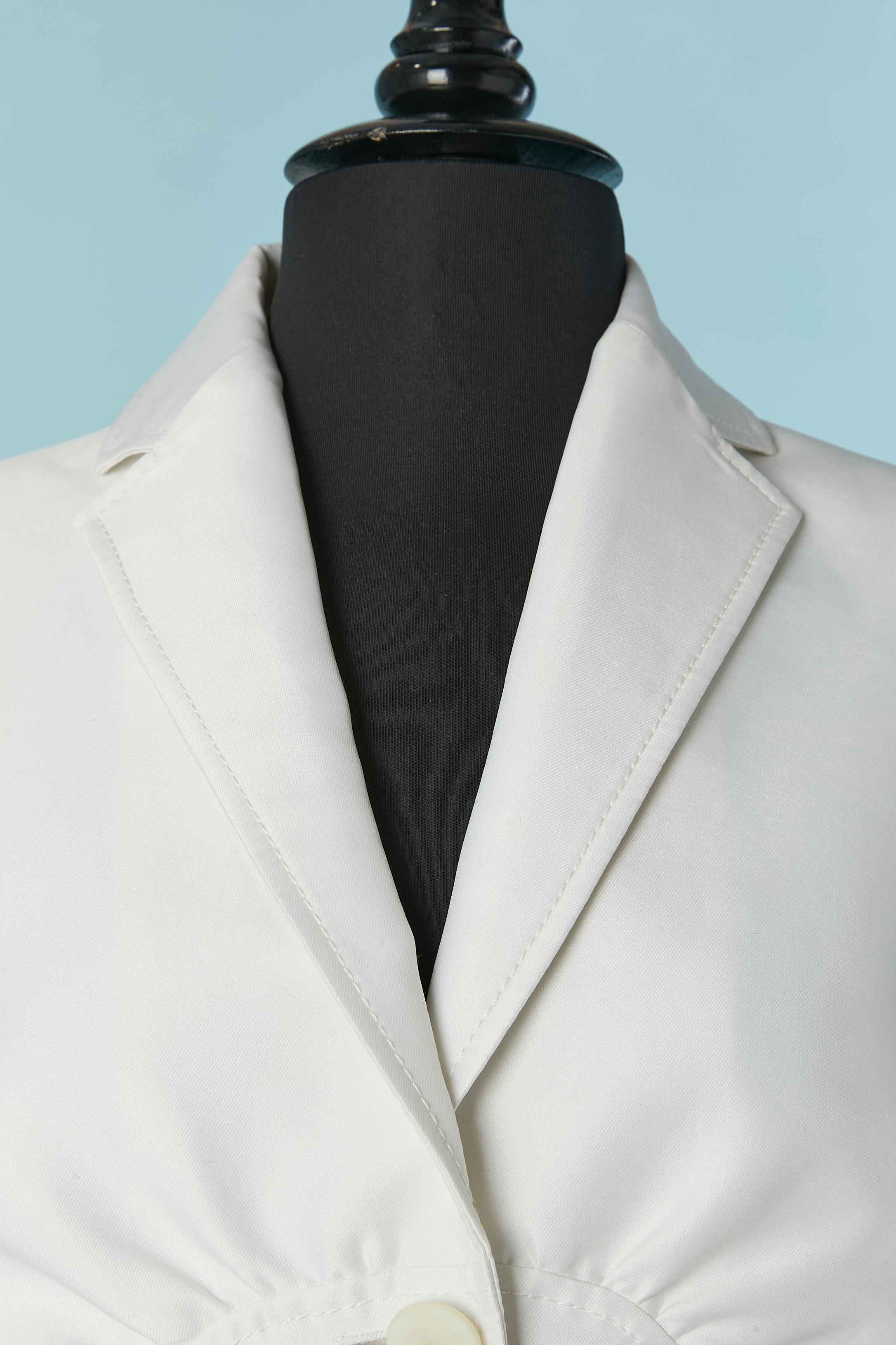 Weißer Rockanzug mit Ausschnitt, Raffungen und Rüschen in der hinteren Mitte. Zusammensetzung des Obermaterials: 81% Polyester, 19% Seide. Zusammensetzung des Futters: 67% Acetat, 33% Polyester. 
Schulterpolster. Markenknopf und Knopfloch in der