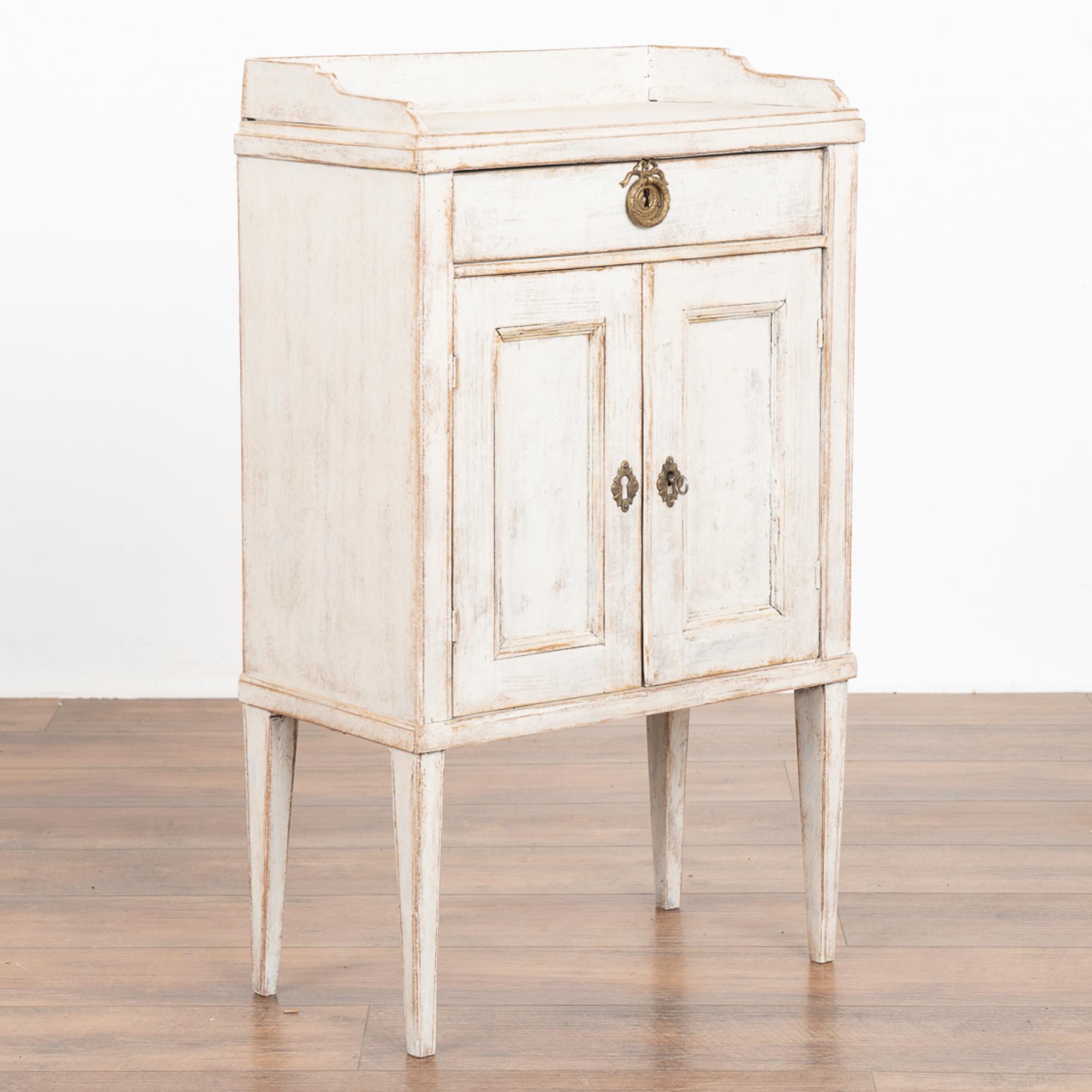 Small Swedish country white painted cabinet standing on tapered legs that may serve also as a nightstand or side table.
A single drawer with brass pull rests above two simple paneled doors.
The lock functions on the right cabinet door and key is