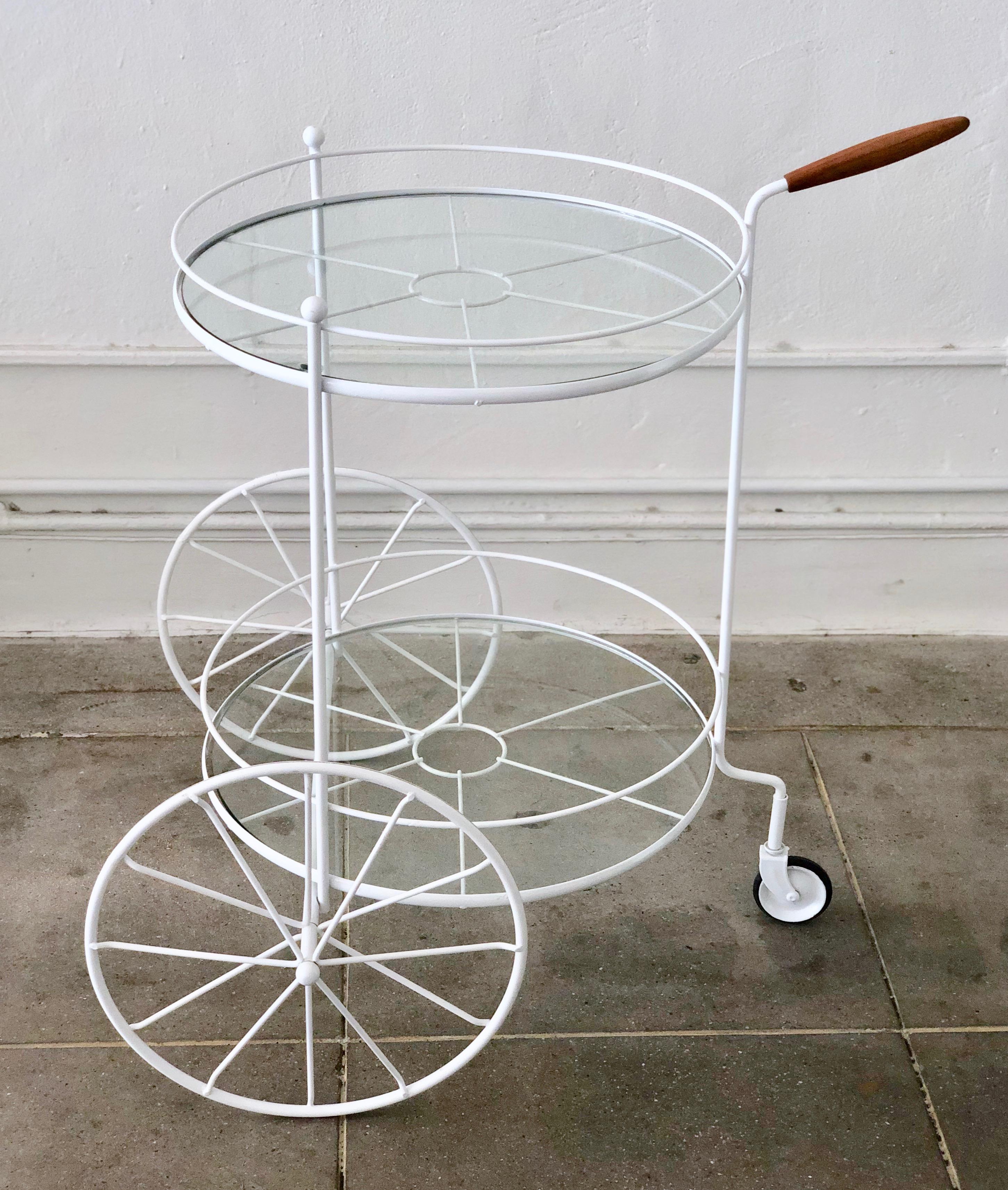 A charming bar cart that is equally at home indoors or outdoors on the patio. This vintage trolley has 2 front large powder coated wheels and a small third wheel in the back and a top handle made of wood. There are two circular pieces of glass
