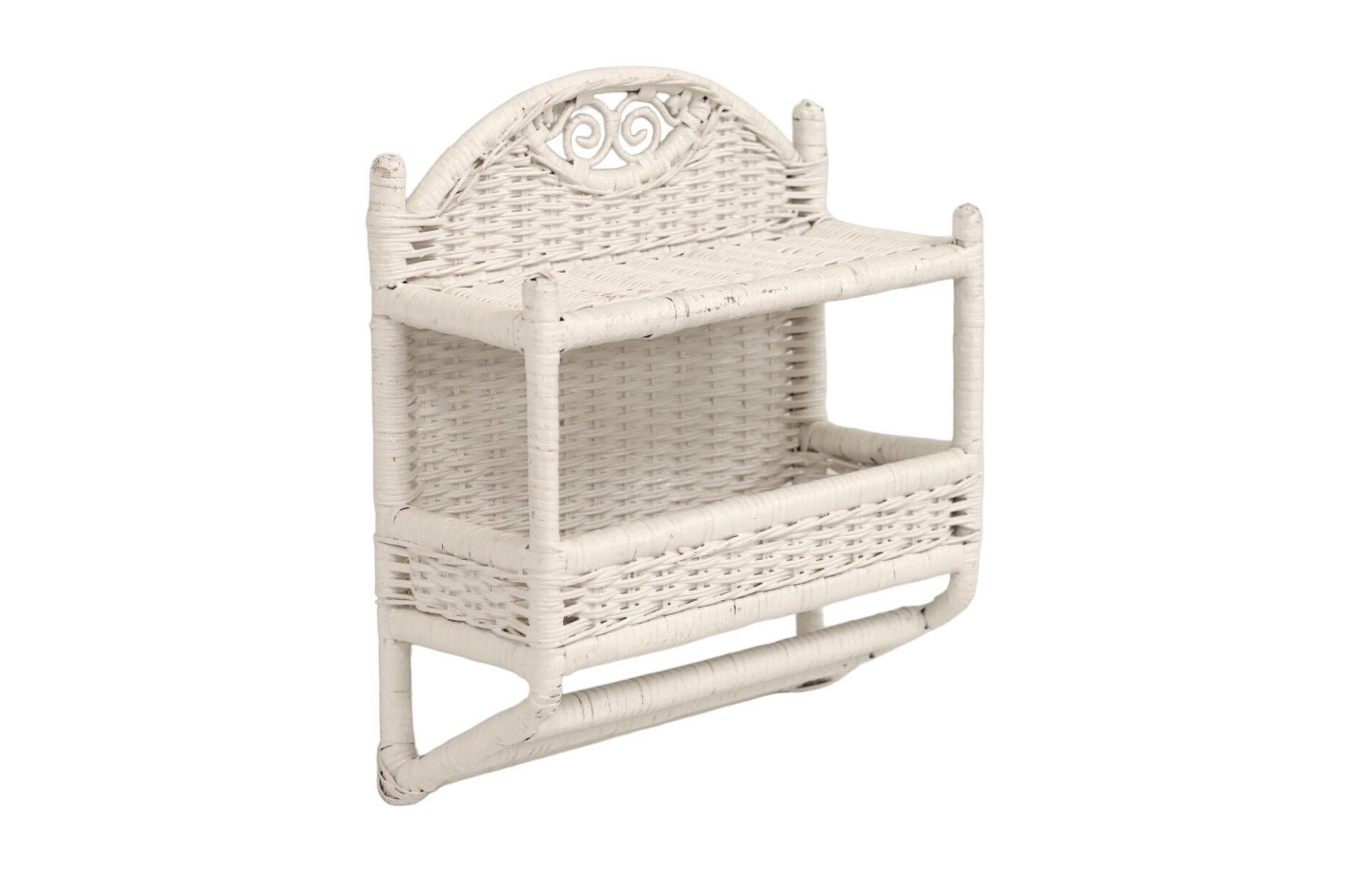 A white bamboo and wicker wall shelf unit. A bamboo frame supports two wicker shelves secured with rattan. Shelves are surmounted with a pierced arch decorated with two scrolls. The lower shelf has small walls and underneath are three rattan bars.