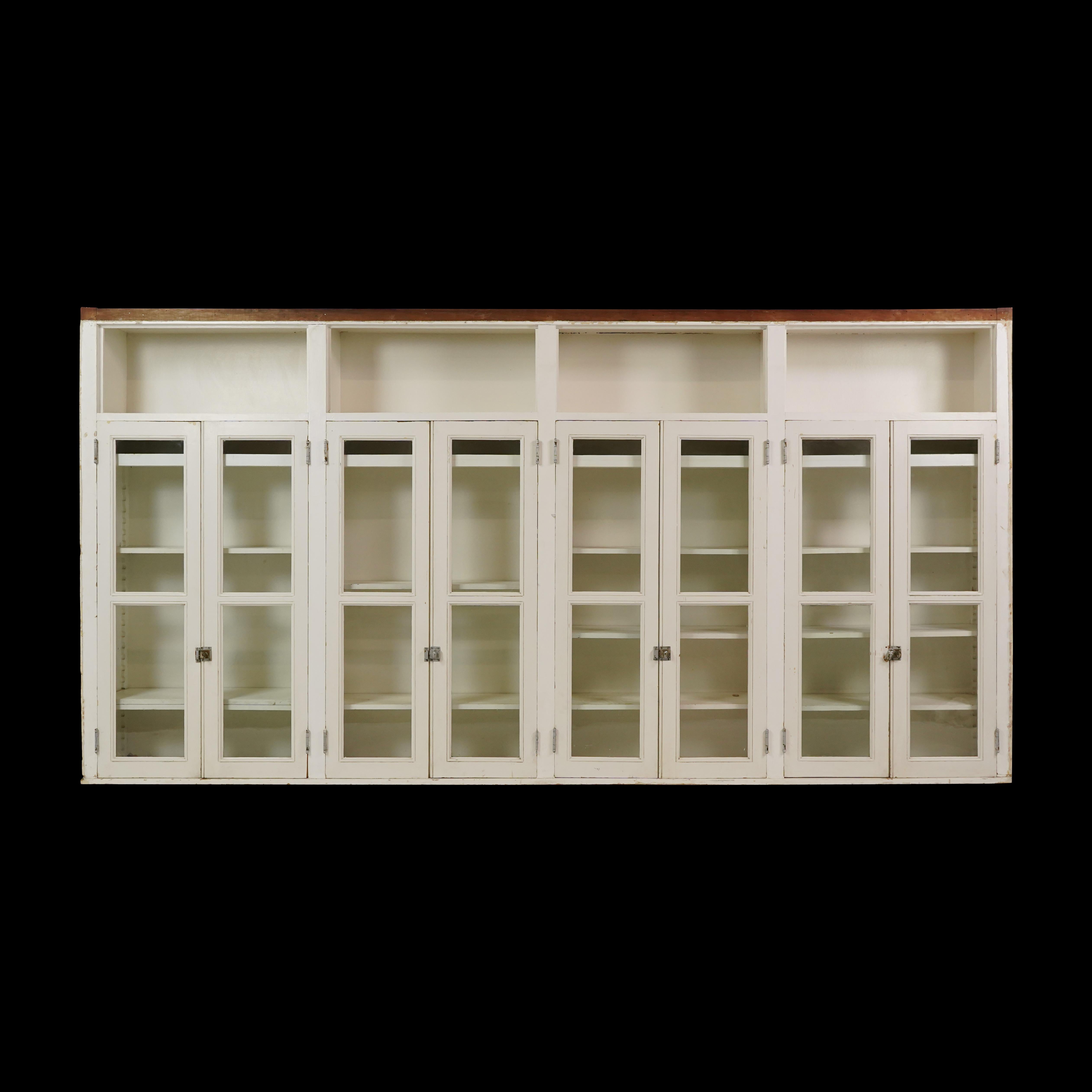 20th Century upper kitchen cabinet unit made of white painted wood and clear glass doors. It features four identical sections with two glass doors, several adjustable shelves, and a top open storage area. The brass cabinet latches look to have been