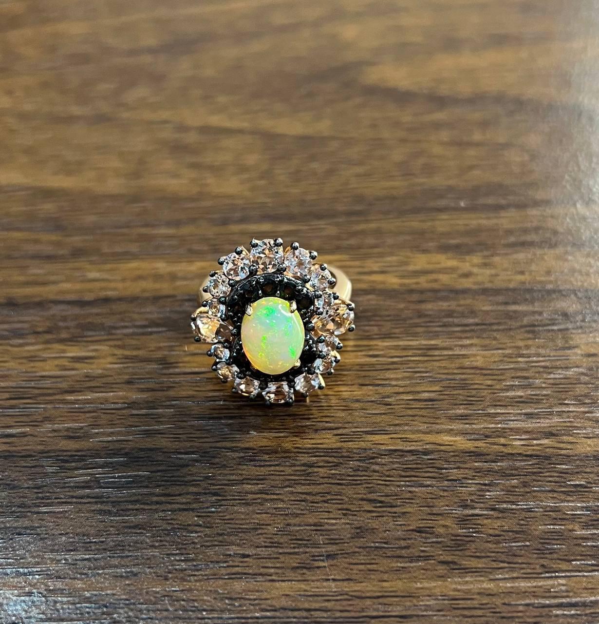 A captivating ring featuring a central opal surrounded by a halo of sparkling black diamonds and peach morganites, set in an intricate design. The opal displays a play of colors, adding depth and allure to the piece. 14K Rose Gold. The surrounding