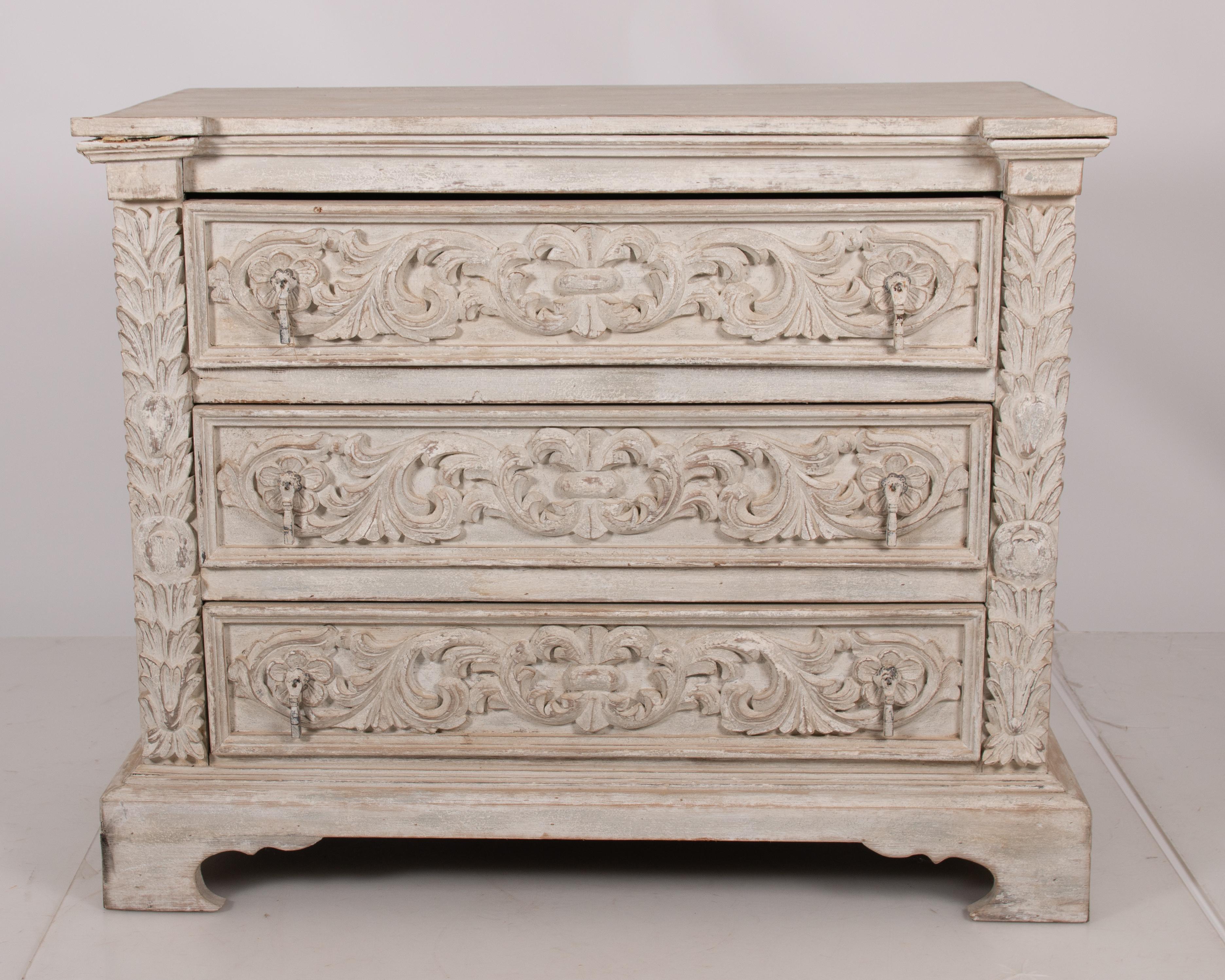 Wonderful English Country style painted wood three-drawer dresser decorated all over with ornate leaf and scroll carvings, made in England circa late 19th century. Exquisitely handcrafted, scrolling acanthus leaf adorn the drawers and square column