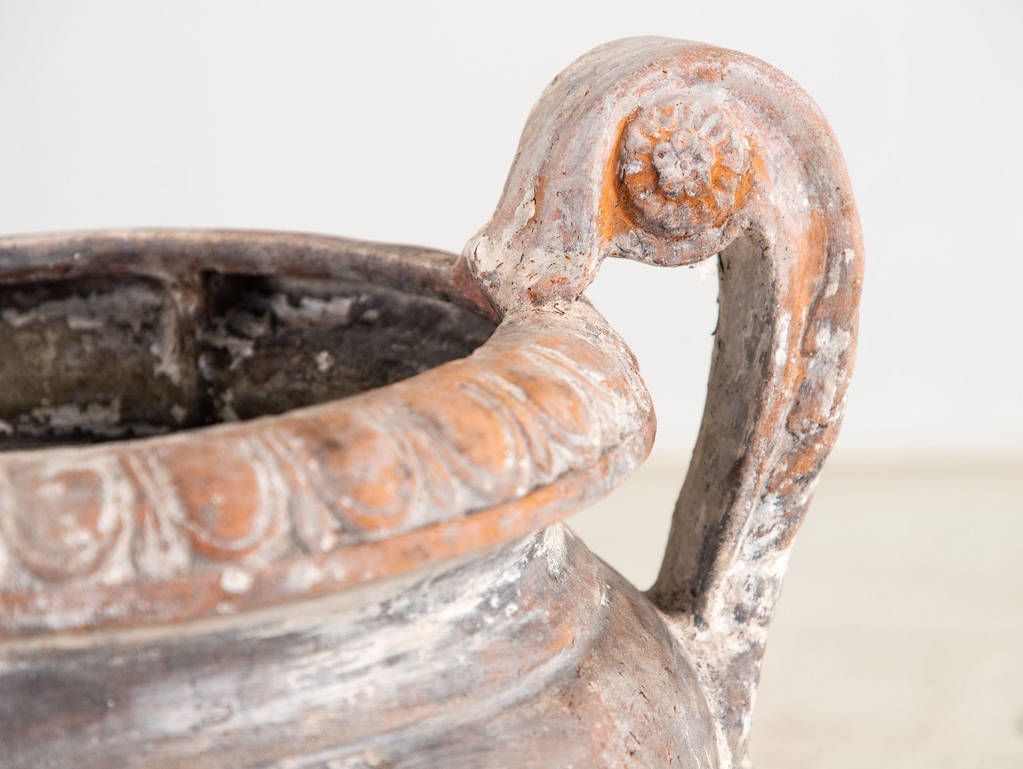 This mid-20th-century Italian terracotta urn features a whitewashed finish, evoking timeless elegance. Its three handles are adorned with delicate acanthus leaf motifs, adding a refined touch. The urn’s lip showcases a traditional egg-and-dart
